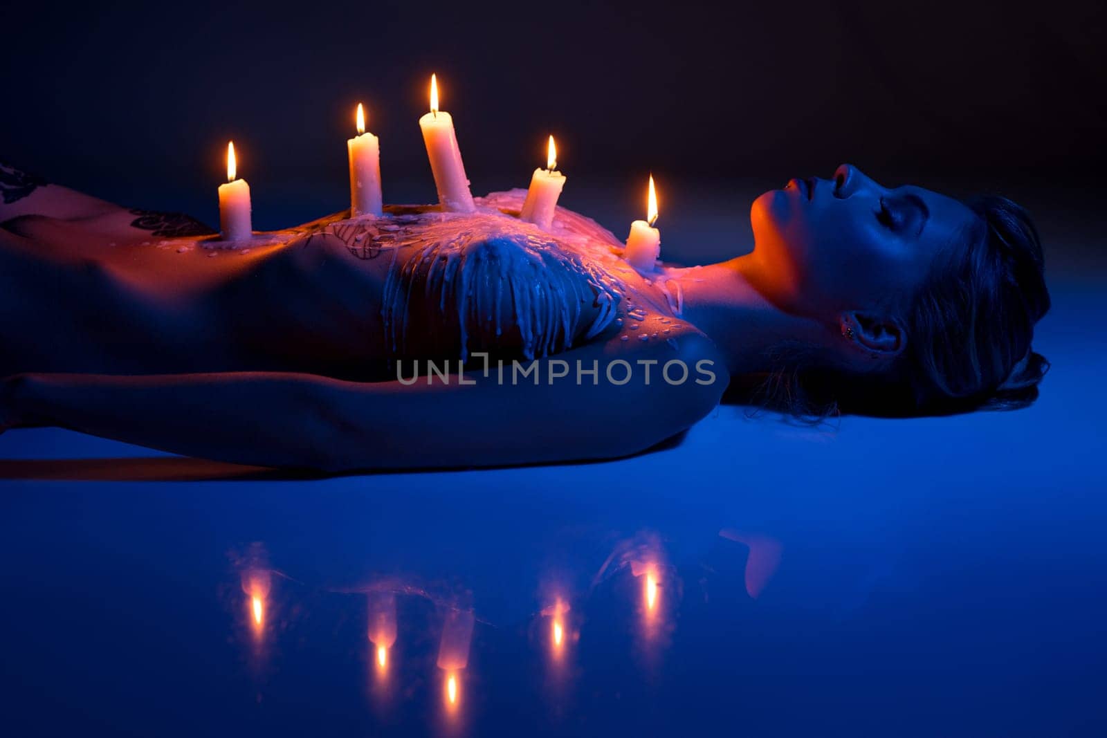 performance of tranquil naked female with burning candles and wax on slim body lying in studio with blue neon light
