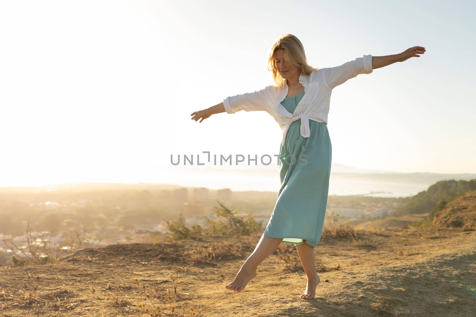 Woman stands on a hilltop at sunset, balancing barefoot with outstretched arms, embracing the freedom and serenity of the moment