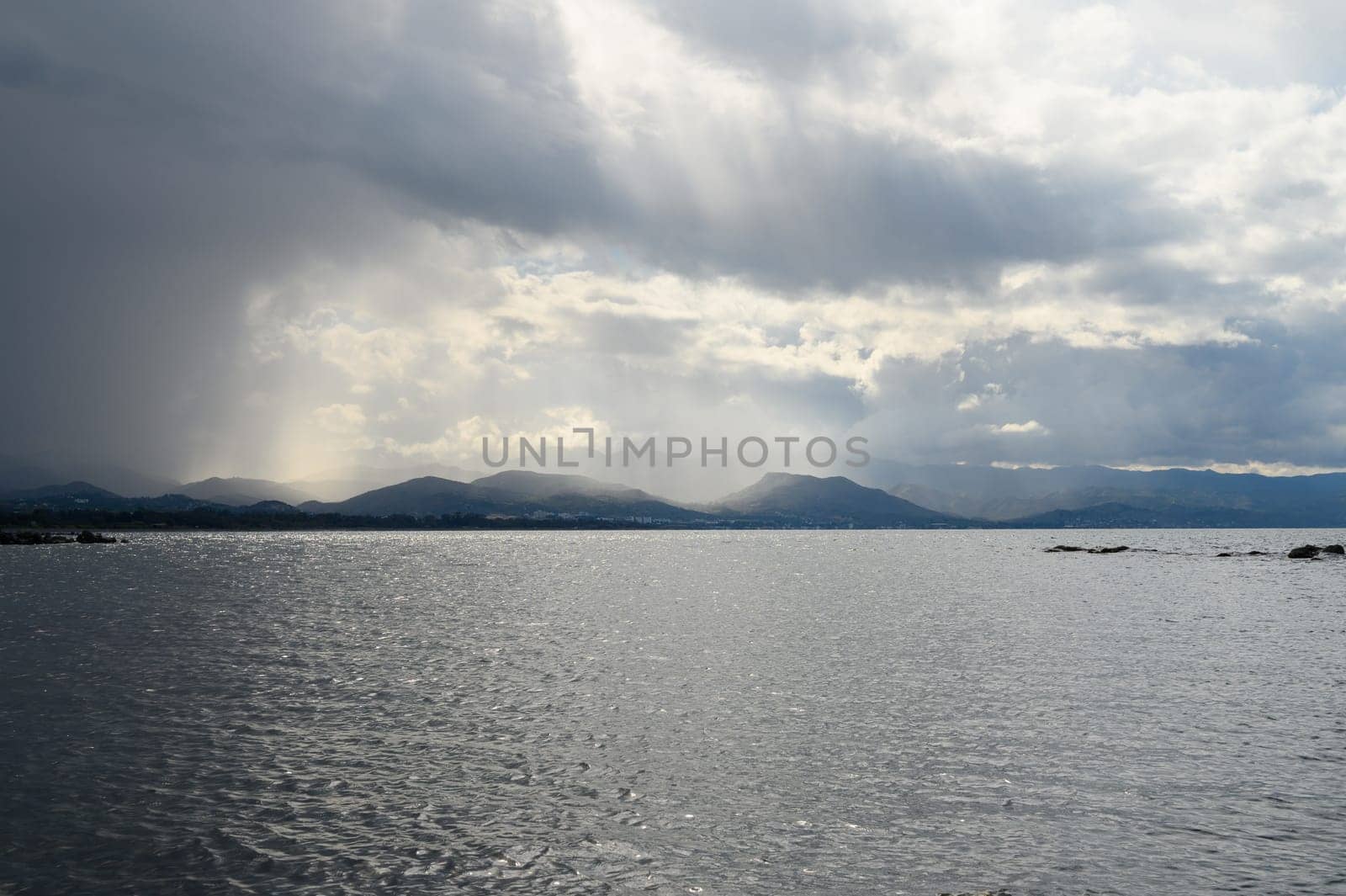 A breathtaking view unfolds as dark clouds loom over a serene body of water. Rays of sunlight break through, illuminating the distant mountains. A moment of nature's splendor captured during midday.