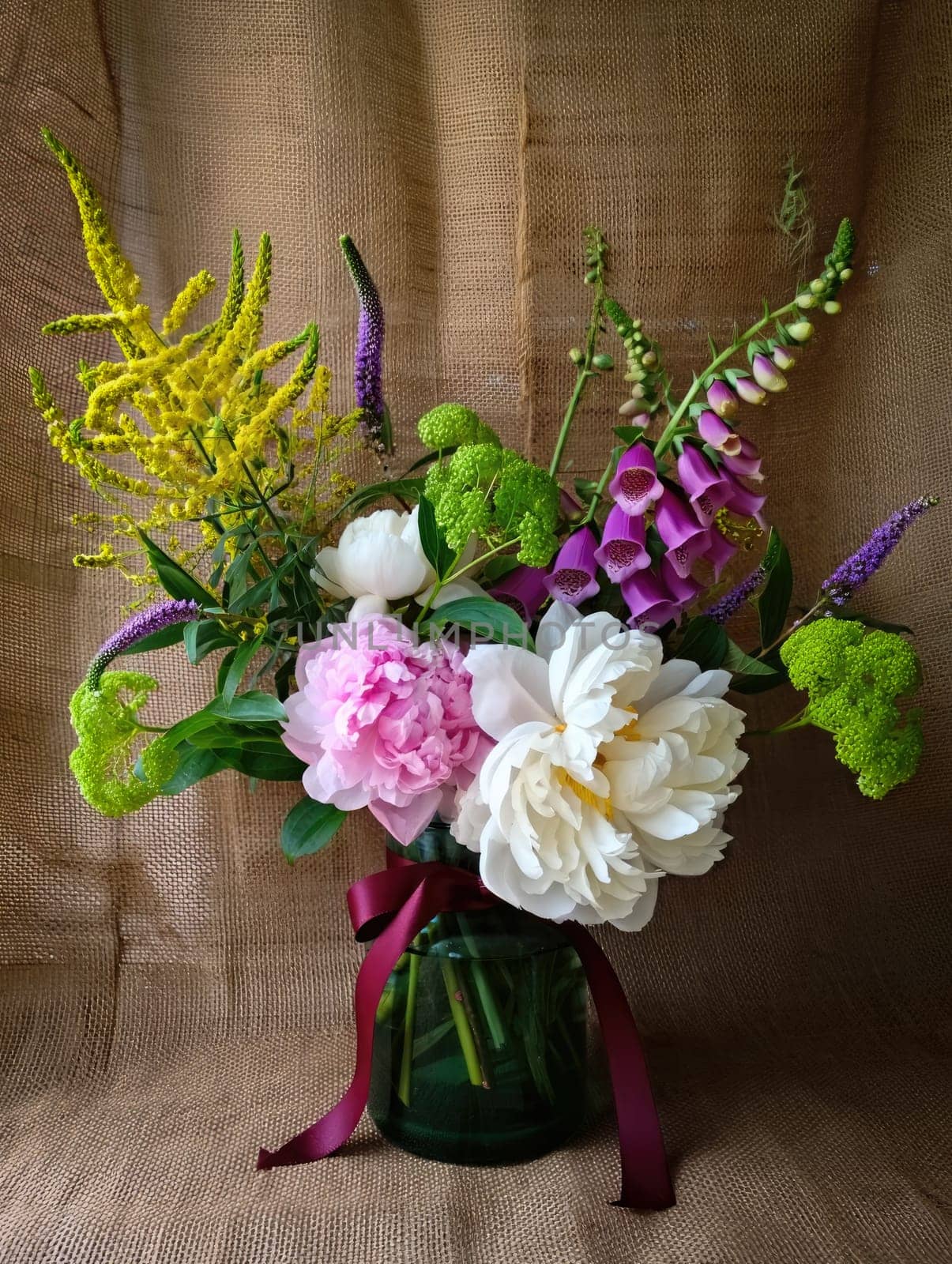 A beautiful bouquet combines peonies, foxgloves, and lush greenery, elegantly arranged in a glass vase. The rustic backdrop enhances the colors and textures of the flowers.
