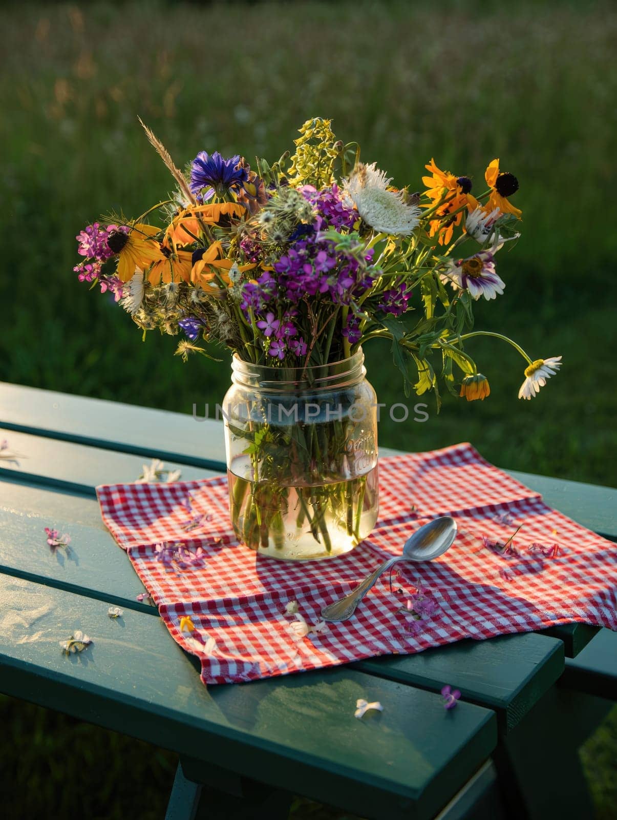 A jar filled with vibrant wildflowers is displayed on a checkered cloth on a green table. The warm golden hour sunlight highlights the flowers and lush greenery in the background.