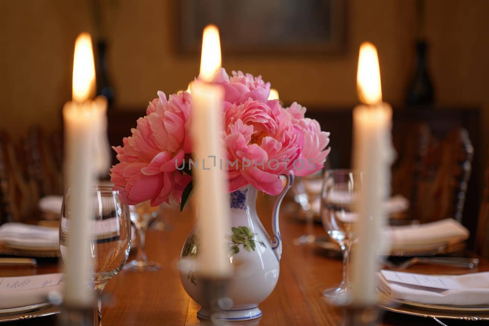 A beautifully arranged dining table features a vase of pink peonies surrounded by lit candles. Glassware is placed neatly, creating a warm and inviting atmosphere for a special gathering.