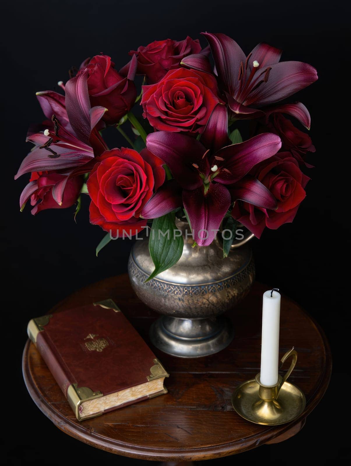 A beautiful arrangement of red roses and lilies showcased in a silver vase on a wooden table. Next to it, a lit candle and an antique book add to the charming ambiance.