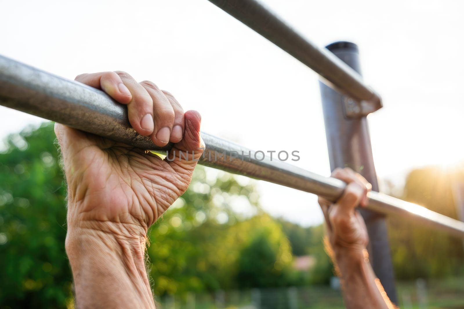 An elderly man is gripping a bar tightly while exercising outdoors in a park. The warm sunlight bathes the scene, highlighting his determination and commitment to fitness.