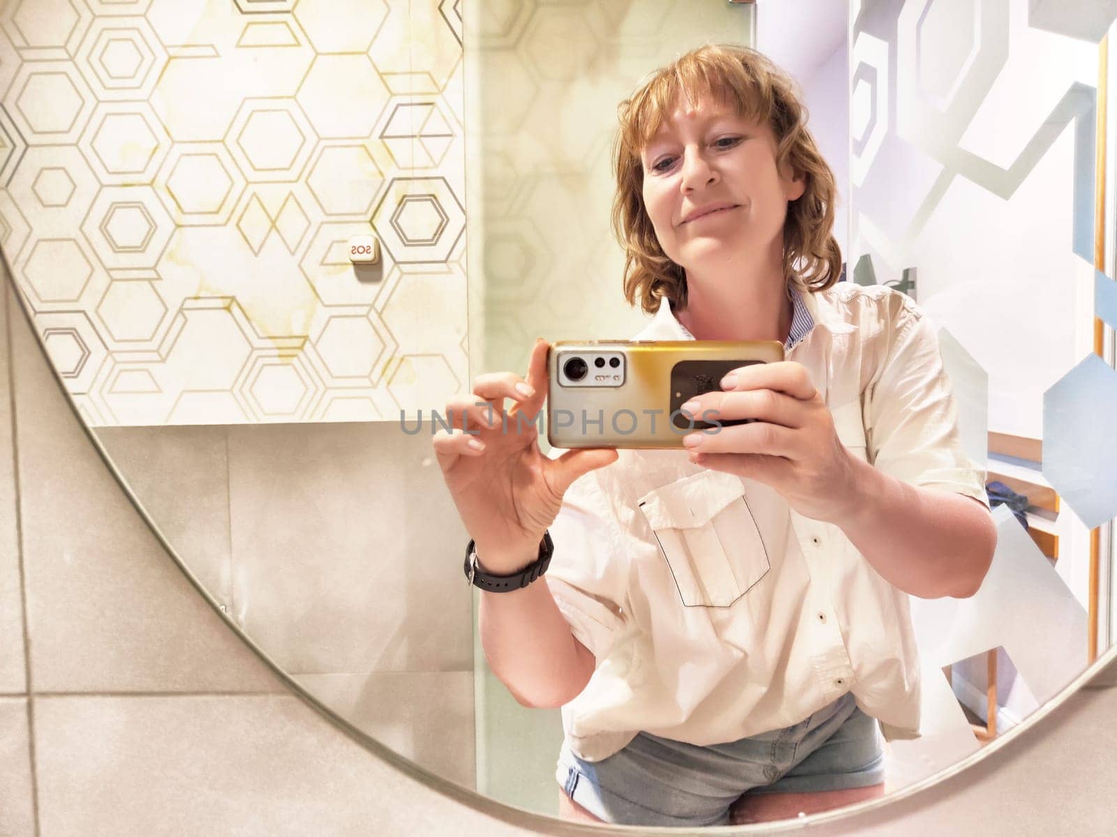 A middle-aged blonde woman smiles while capturing a reflection selfie in a stylish restroom, showcasing her blogging spirit and lifestyle.