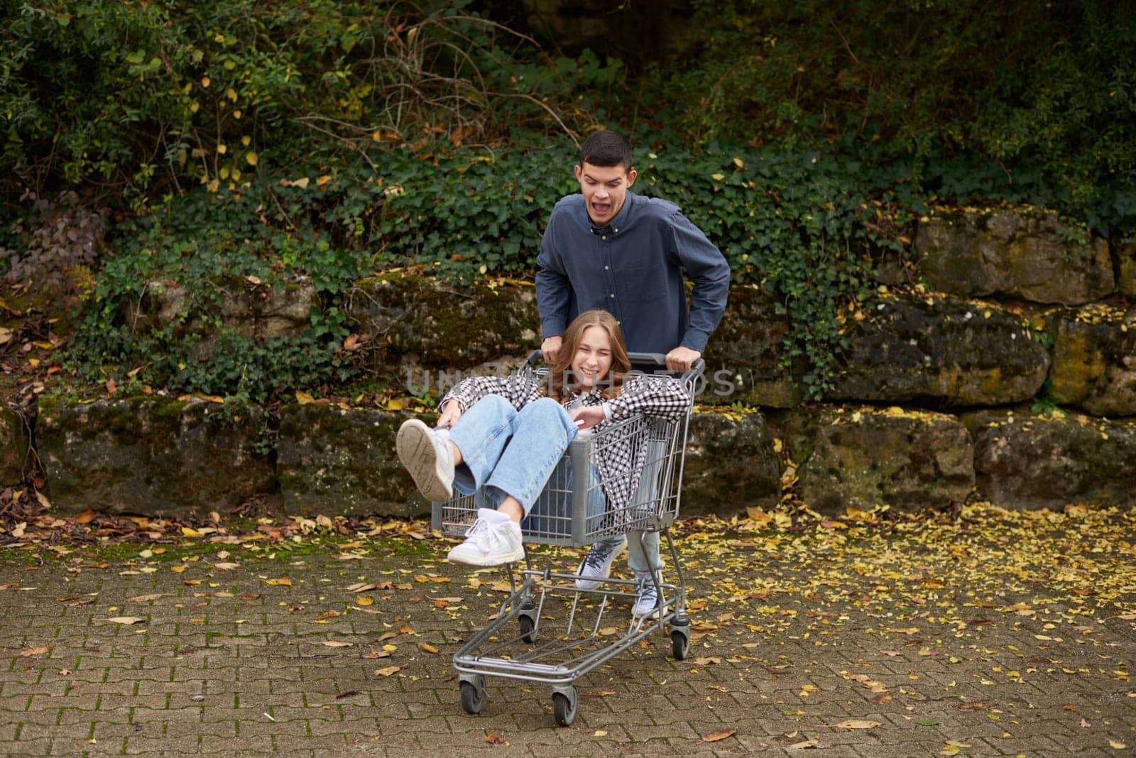A cheerful young couple enjoys an adventurous ride with a shopping cart on an outdoor paved path, surrounded by autumn foliage and greenery. The young woman, dressed in a trendy checkered flannel shirt and light blue jeans, sits inside the cart with a bright smile, while the man, wearing a navy blue jacket and jeans, pushes the cart with playful enthusiasm. The scene is filled with energy, joy, and spontaneity, making it ideal for lifestyle photography, relationship themes, autumn fashion campaigns, and advertising related to youth, fun, and carefree moments. The natural warm lighting and seasonal tones enhance the cozy and nostalgic atmosphere, making this image a great choice for social media content, blogs, and promotional materials focused on friendship, romance, and outdoor activities.