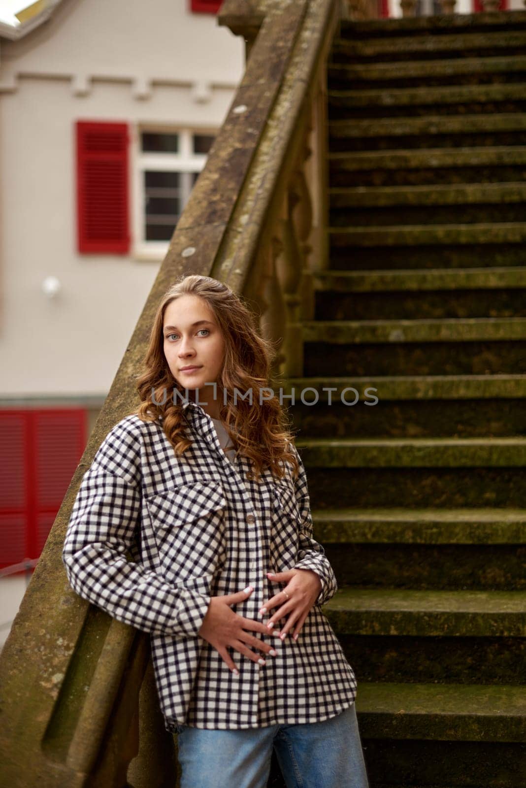 This dynamic and lively image captures a beautiful young woman flipping her hair in a carefree manner while standing in a meticulously maintained garden, possibly located in a charming European village. She is dressed casually in a white long-sleeve crop top, blue jeans, and white sneakers, exuding a modern, relaxed style. Her casual attire, paired with the confident gesture of flipping her hair, adds a sense of movement and vitality to the scene. The background features vibrant pink hydrangeas and traditional half-timbered houses with green shutters and steep roofs, characteristic of European architecture. The lush greenery and colorful flowers create a tranquil yet energetic atmosphere, making this photo perfect for themes such as lifestyle, travel, seasonal fashion, natural beauty, and rural European culture. The intentionally blurred face maintains the subject's privacy, adding an element of mystery and anonymity.Beautiful Young Woman Flipping Hair in a Lush European Garden with Pink Hydrangeas and Traditional Houses