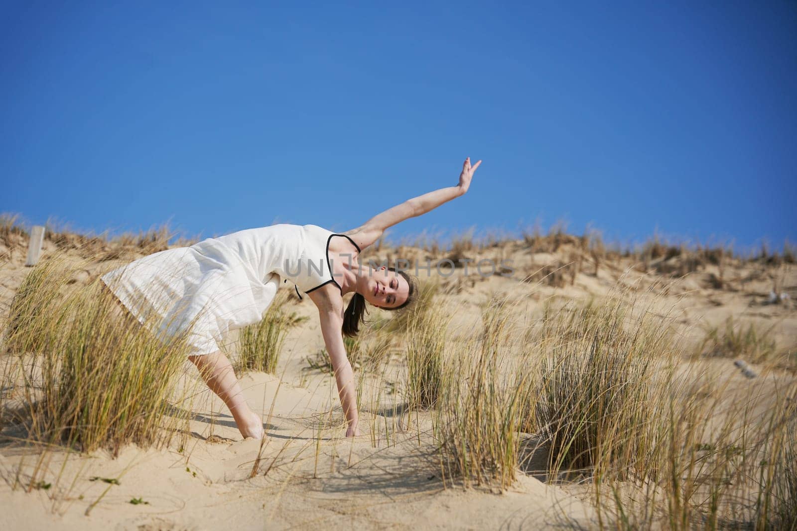 A barefoot woman in a white summer dress performs a graceful pose on a sand dune. The bright blue sky and surrounding dry beach grass create a calm and artistic atmosphere.