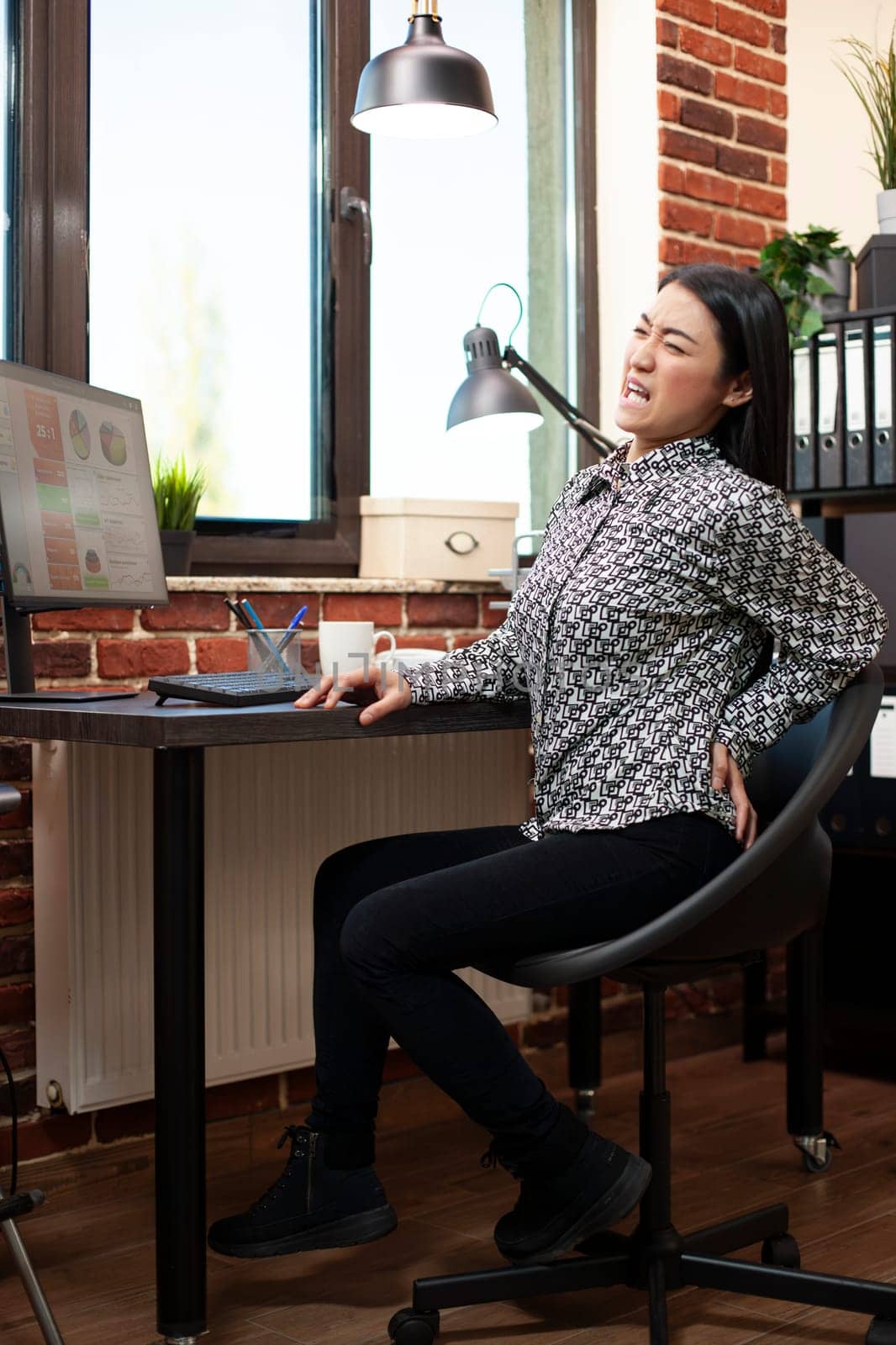 Asian businesswoman sitting at desk and holding her lower back, feeling discomfort at work. Female employee struggling with backpain, visibly uncomfortable on her chair in brick wall office.