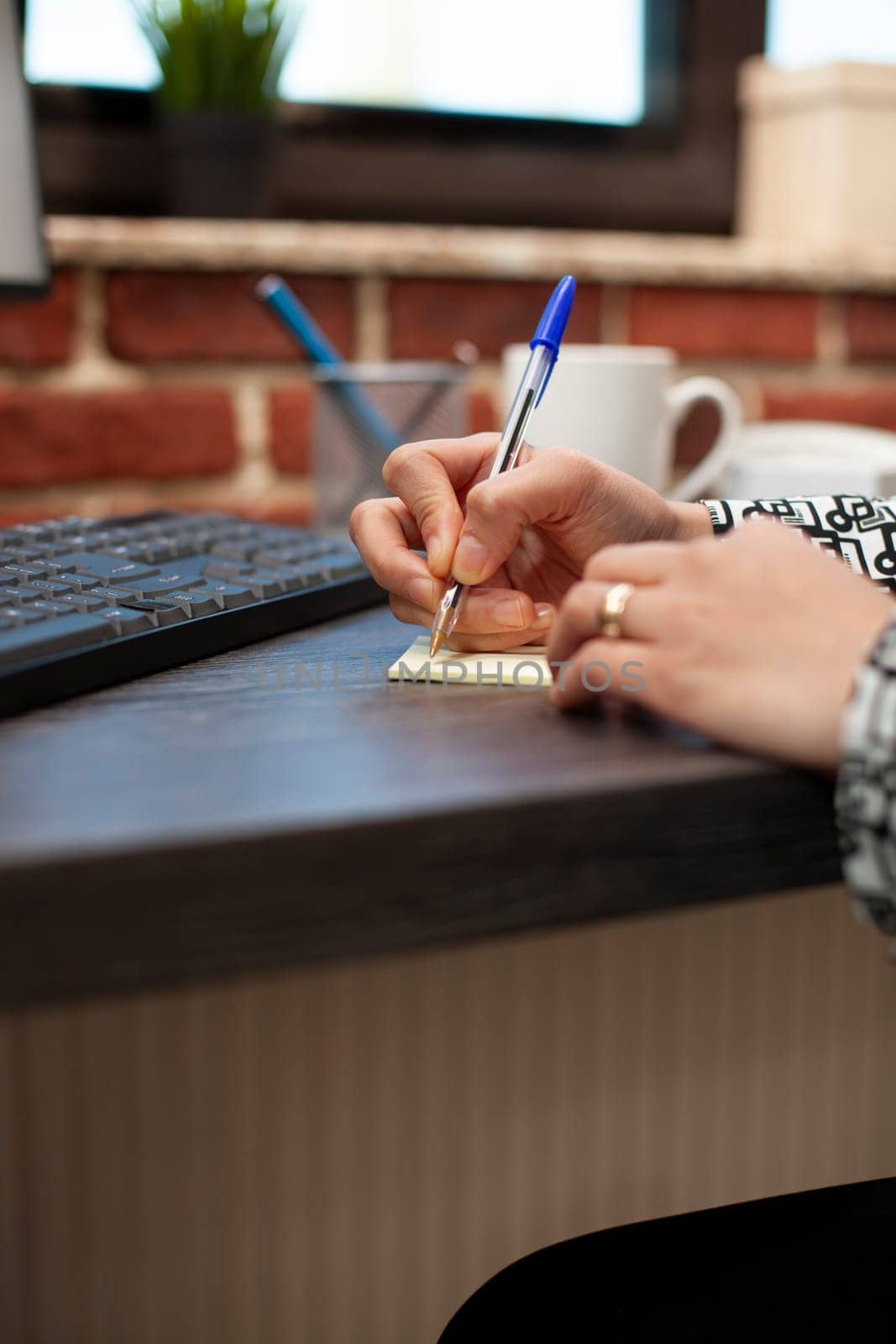 Closeup of business person writing on sticky note on office desk, planning work schedule. Selective focus on hands of female employee using a pen, noting down company strategy ideas on table.