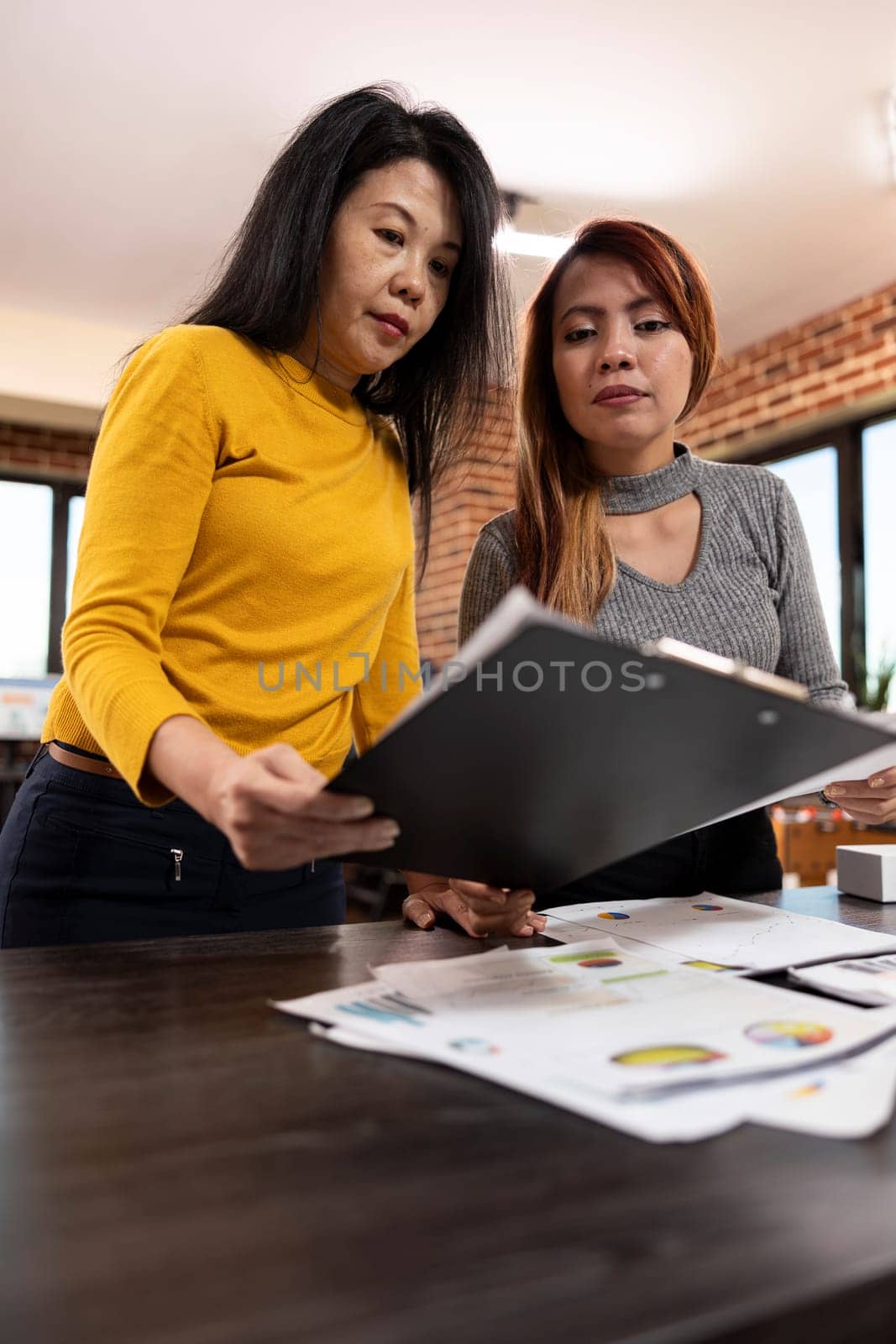 Closeup of female coworkers standing and looking at a clipboard in brick wall workplace. Asian businesswomen reviewing together startup company documents and collaborating near office desk.