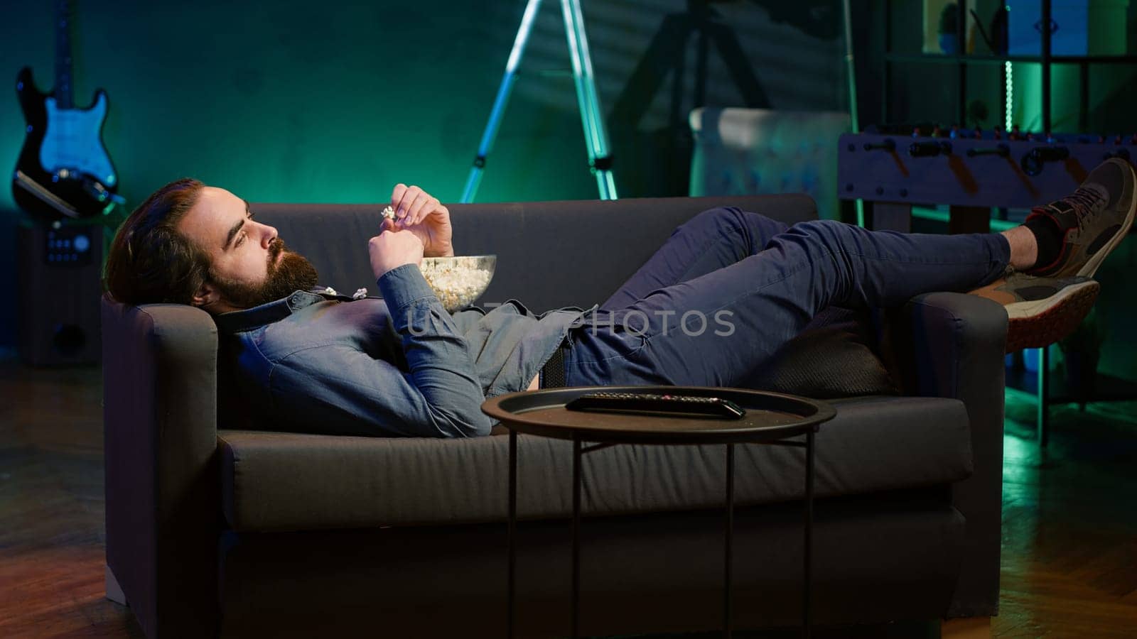 Man procrastinating at home, laying down on couch, munching on snacks, watching TV. Bored person living sedentary lifestyle, lounging on couch, wasting time watching TV, eating popcorn, camera A