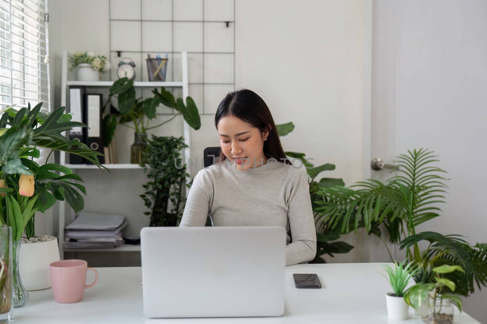 A woman sits intently at her desk, using her laptop to complete tasks, highlighting the productivity of a serene work environment filled with plants.