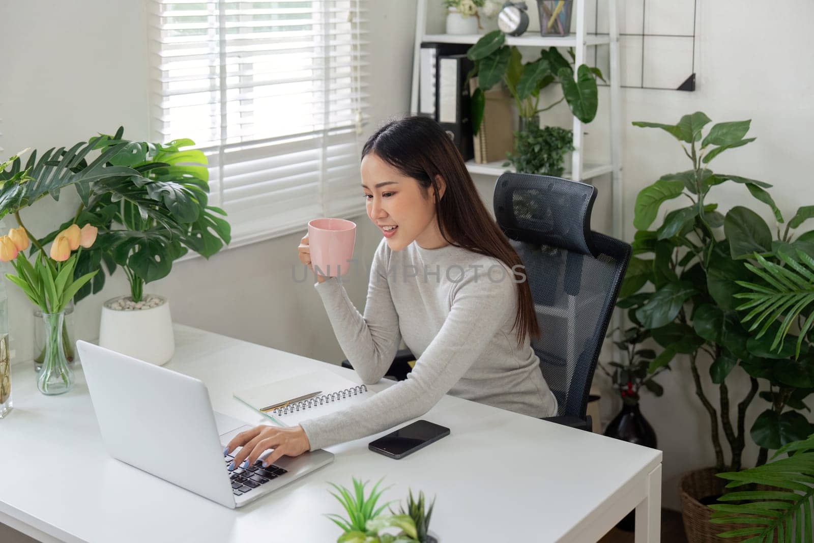 A young woman sits comfortably in her home office, sipping a coffee and engaging with her laptop, blending relaxation with productivity.