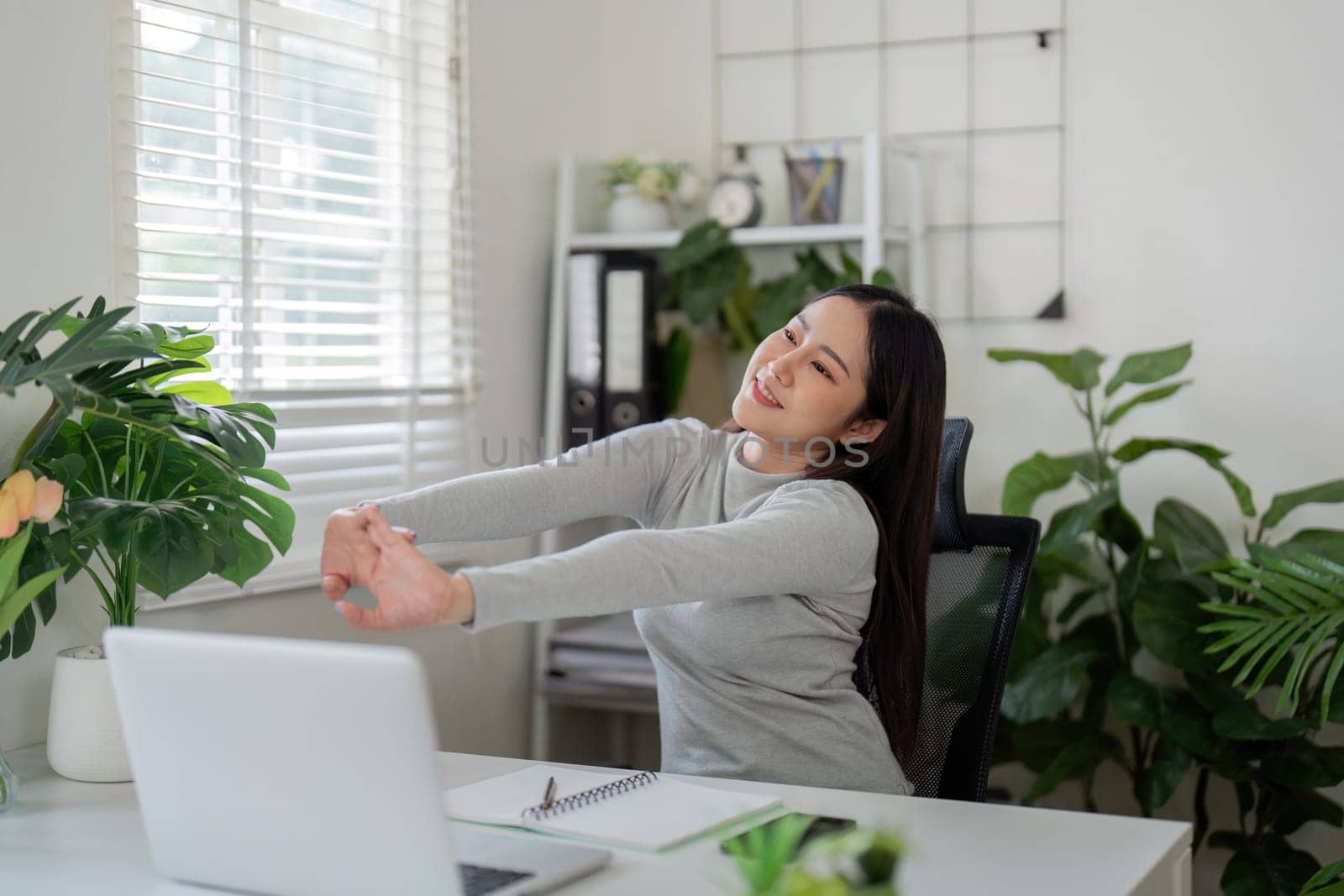 In her serene workspace, a young woman does a gentle stretch, promoting wellness and relaxation while working from home.