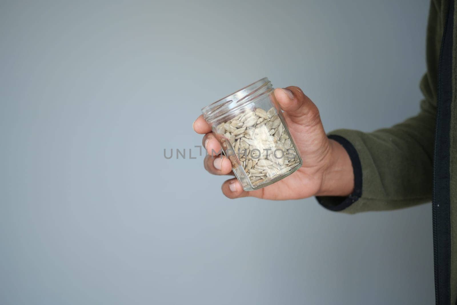 A person holds a small jar containing various seeds in front of a plain wall.