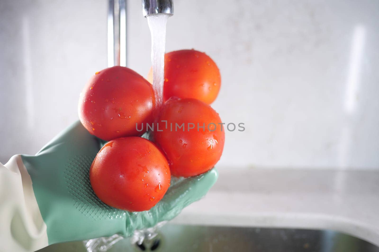 Hands in gloves rinse red tomatoes under running water in the kitchen.