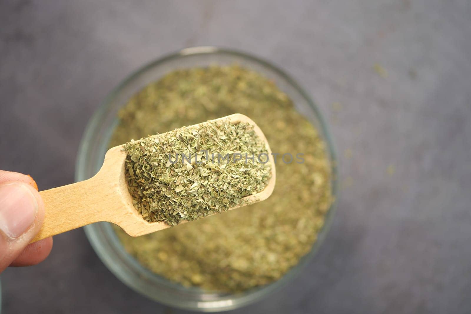 A wooden scoop holds green herbs above a glass container filled with more.