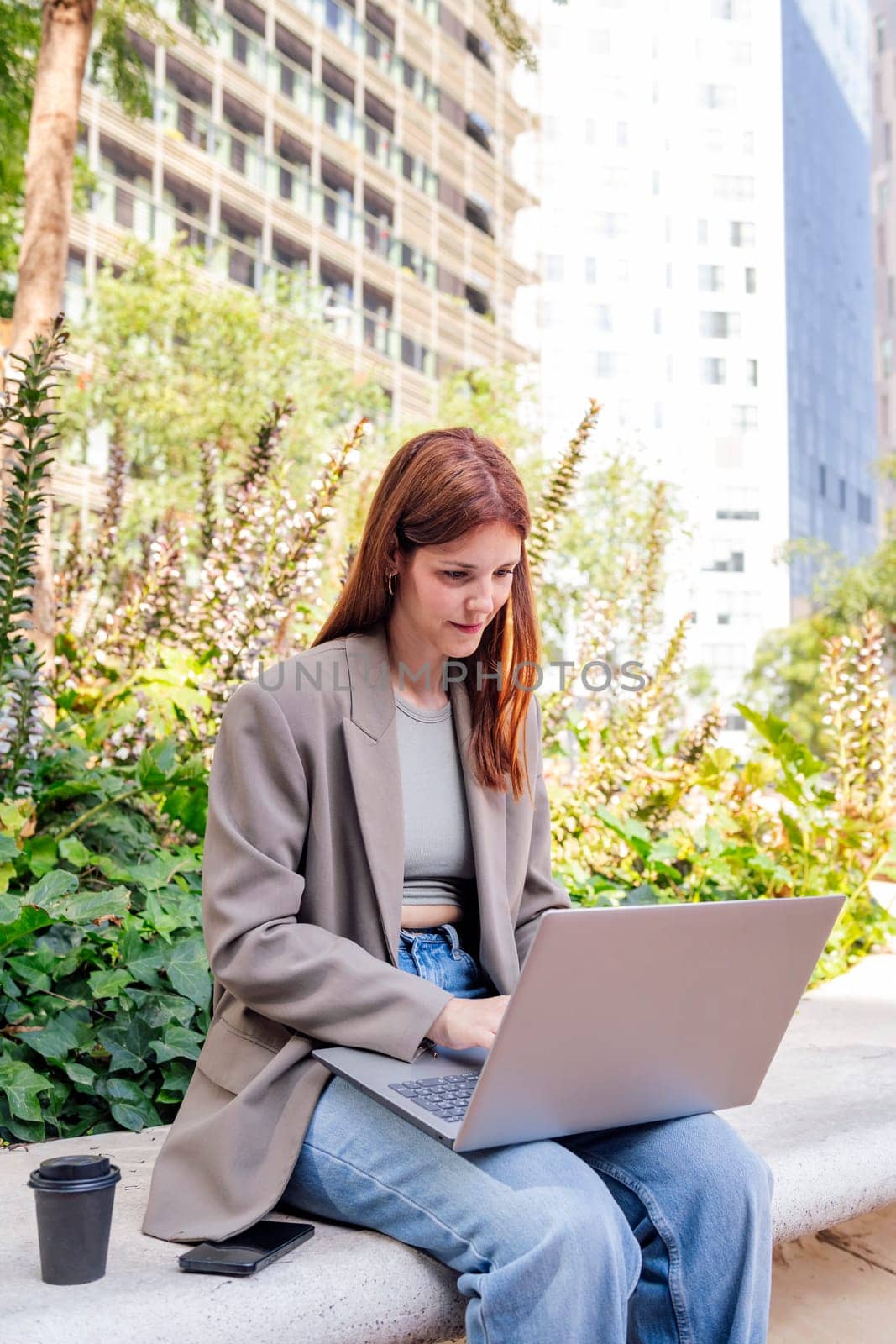 smiling young woman using a laptop sitting on a bench of a park near of office buildings, concept of business and technology for remote work