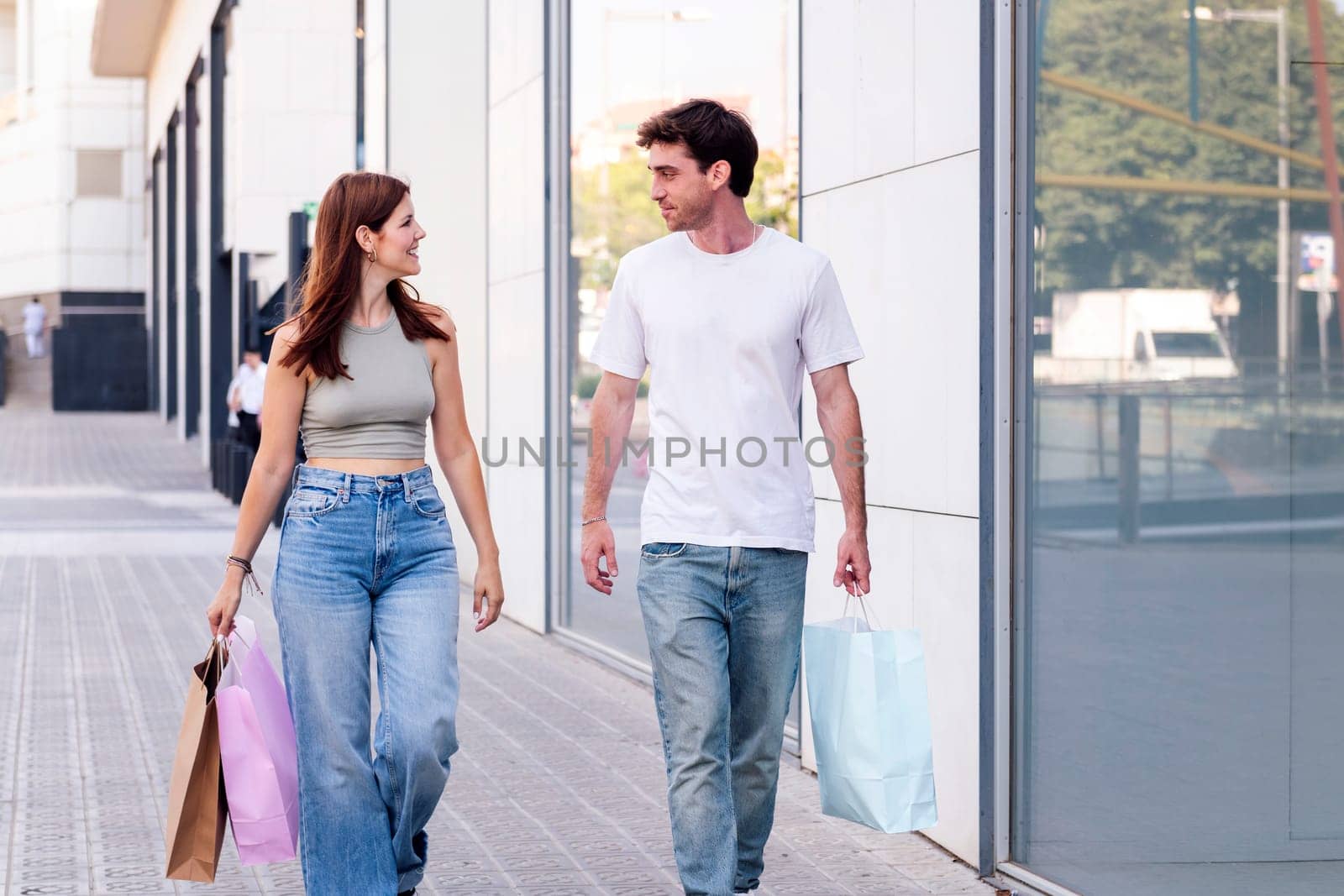 young adult man and a woman in casual clothes walking down a sidewalk near of a mall carrying shopping bags, concept of couple and urban lifestyle, copyspace for text