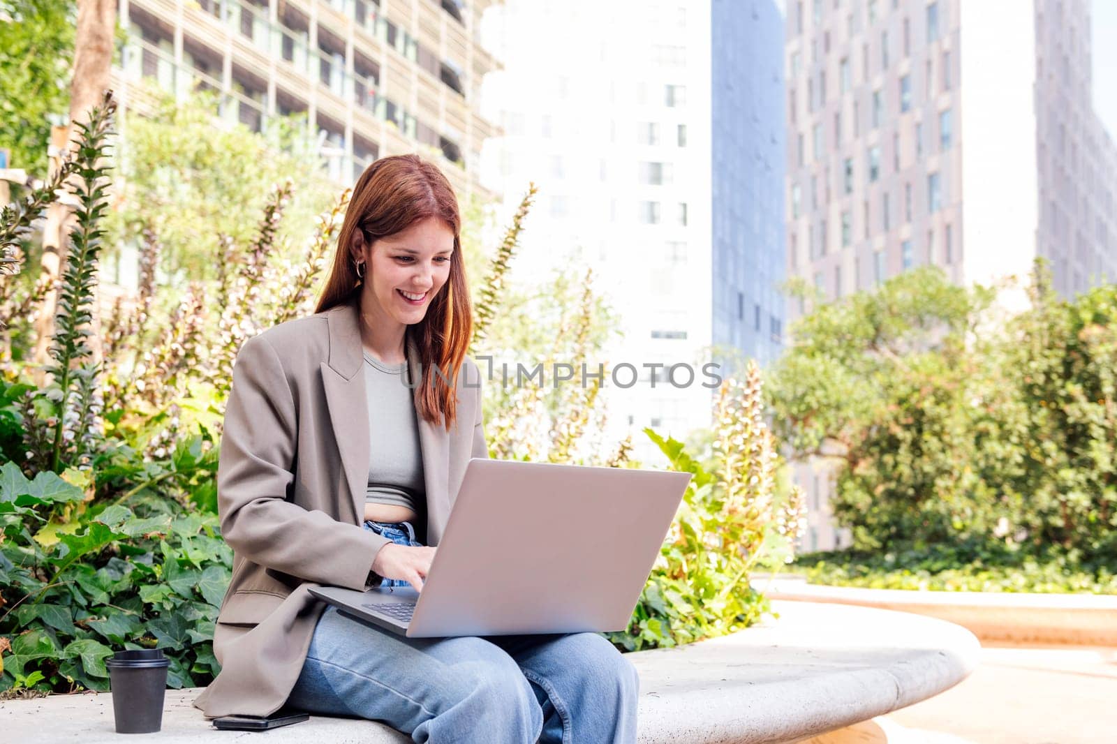 smiling young woman using a laptop sitting on a bench of a park near of office buildings, concept of business and technology for remote work