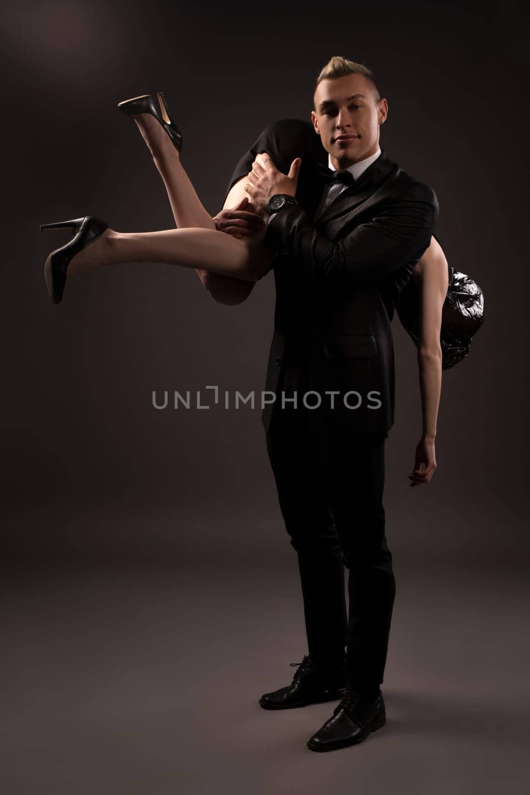Full length of man in suit lifting woman with bag on head while playing BDSM game in dark studio and looking at camera