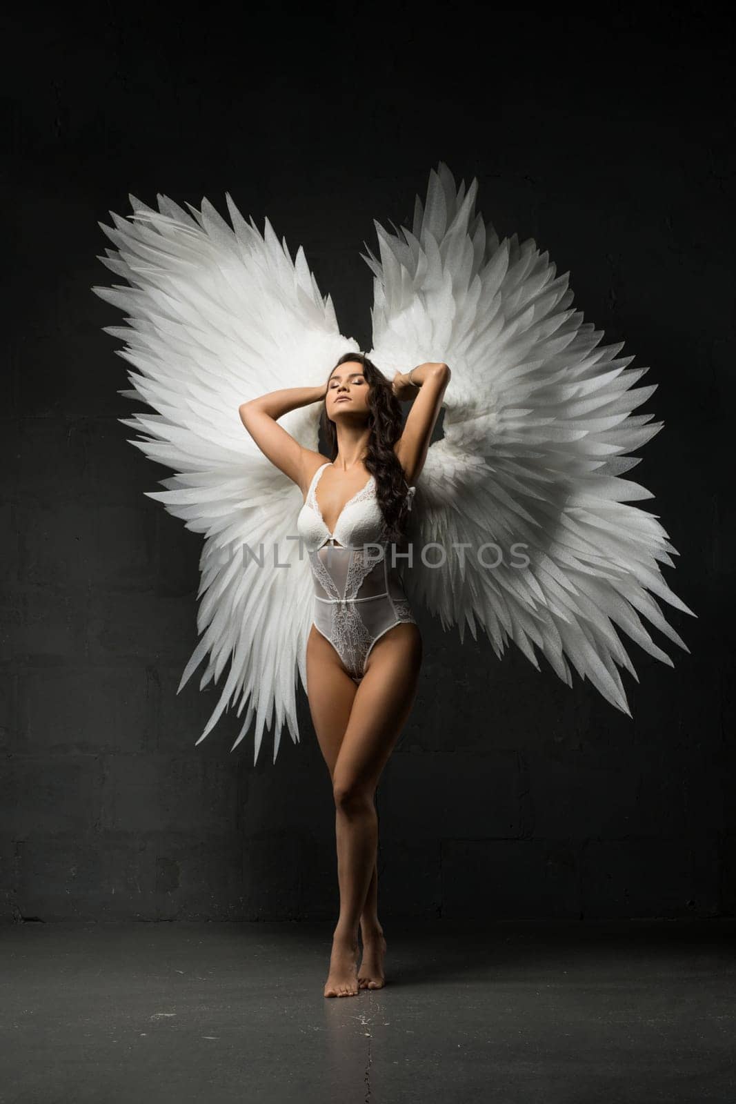 Full body of gorgeous female model wearing white bodysuit and angel wings standing with crossed legs on tiptoes with eyes closed and hands behind head