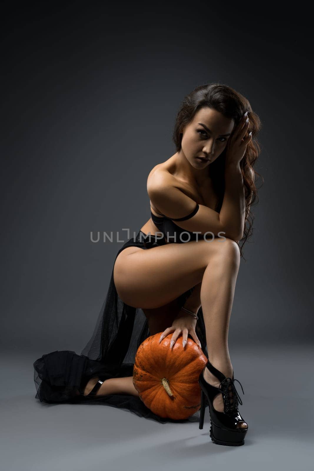 Full body of seductive female model wearing black underwear and long skirt with high heels leaning on pumpkin and looking at camera with sensual glance