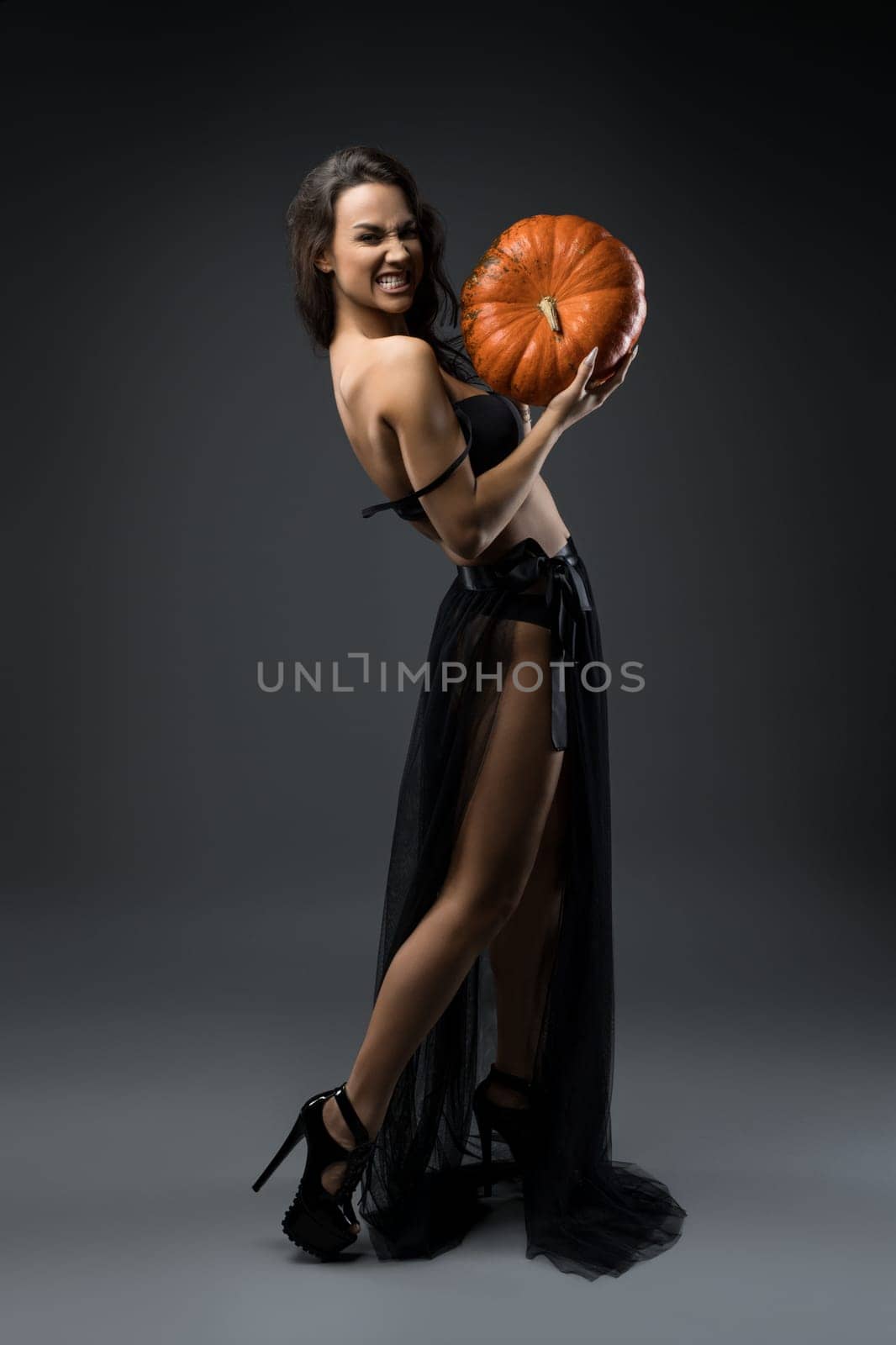 Full body side view of slim female model wearing black maxi skirt and lingerie standing with orange pumpkin and looking at camera with angry grin