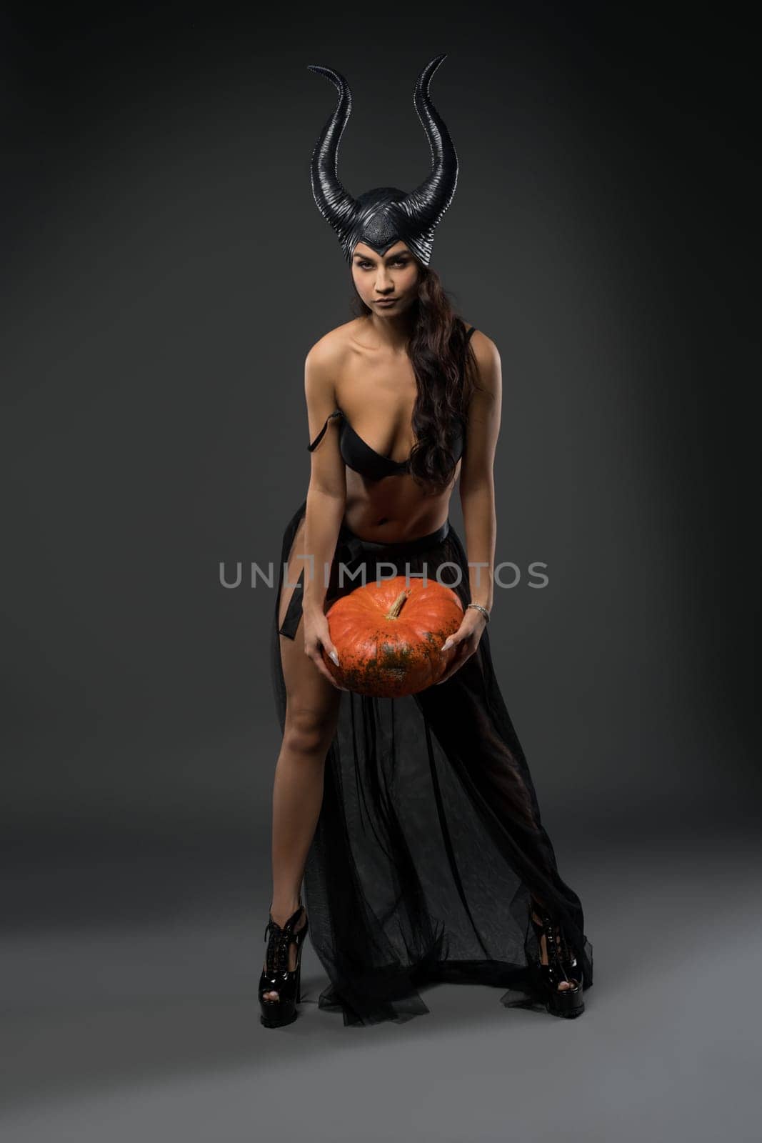 Full body of smiling female witch model wearing provocative black devil fairy with horns costume standing with pumpkin in hands
