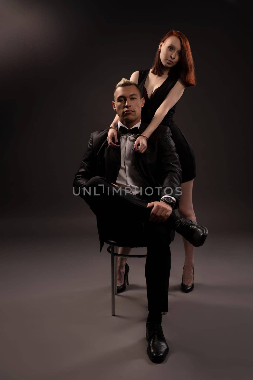 couple portrait of woman in dress dominant man with handcuffs sitting on chair while enjoying foreplay during BDSM session