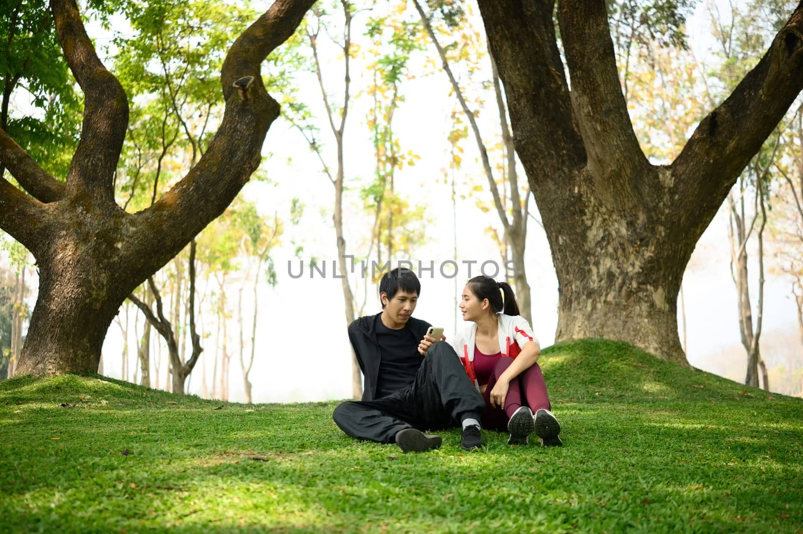 Happy couple sitting on green grass in a peaceful park and looking at smartphone.