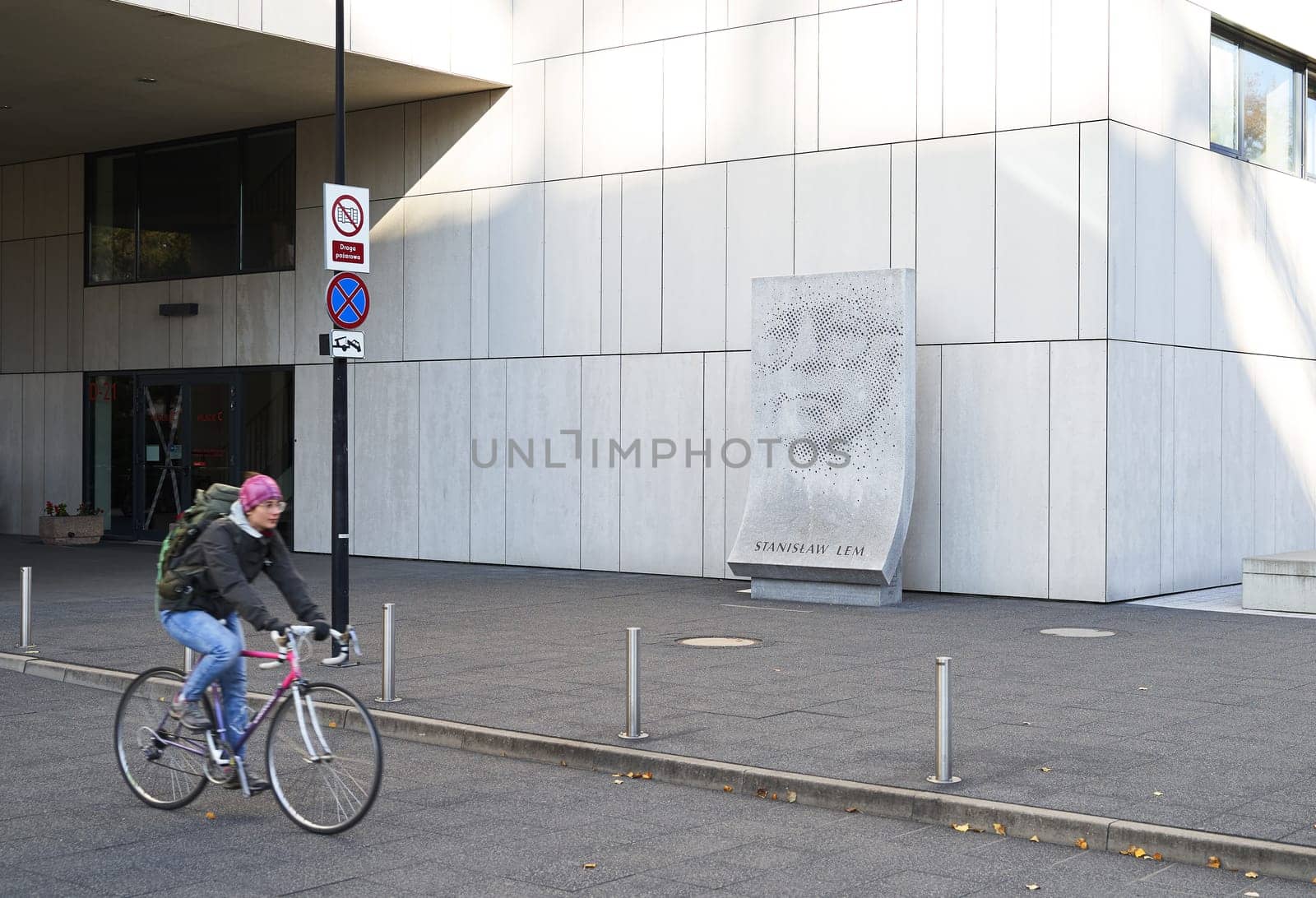 Wroclaw, Poland - October 24, 2024: Bicycle driver passes the sculpture of Stanislaw Lem located next to the Library building of Wroclaw University of Science and Technology. The three-metre-high work, made of Strzegom granite and designed by Grzegorz Niemyjski, PhD, professor at the Academy of Fine Arts in Wroclaw, depicts the writer's face in the form of small holes drilled into the surface of a block of rock.