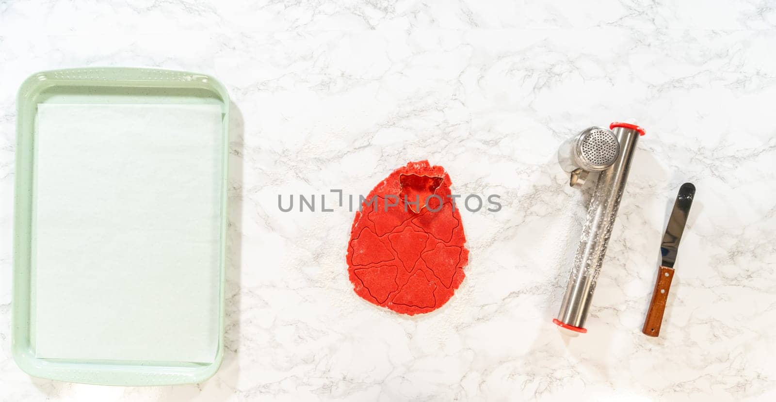 On a marble countertop, a smooth red ball of gingerbread dough is ready to be rolled. Next to the dough, a rolling pin, flour sifter, and offset spatula are arranged, creating a clean and organized baking setup for making Red Gingerbread Cookies.
