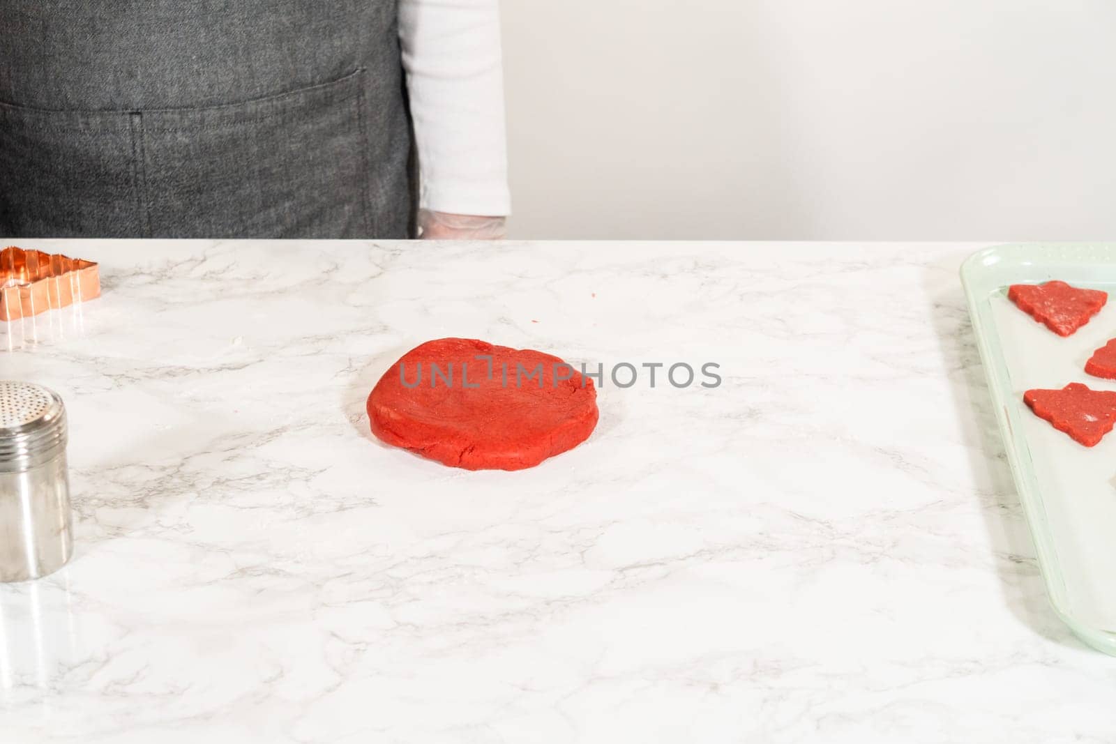 Small, tree-shaped pieces of red gingerbread dough are arranged on a marble countertop, ready for transfer to the baking tray. These festive shapes are part of the Red Gingerbread Cookies recipe for the holiday season.
