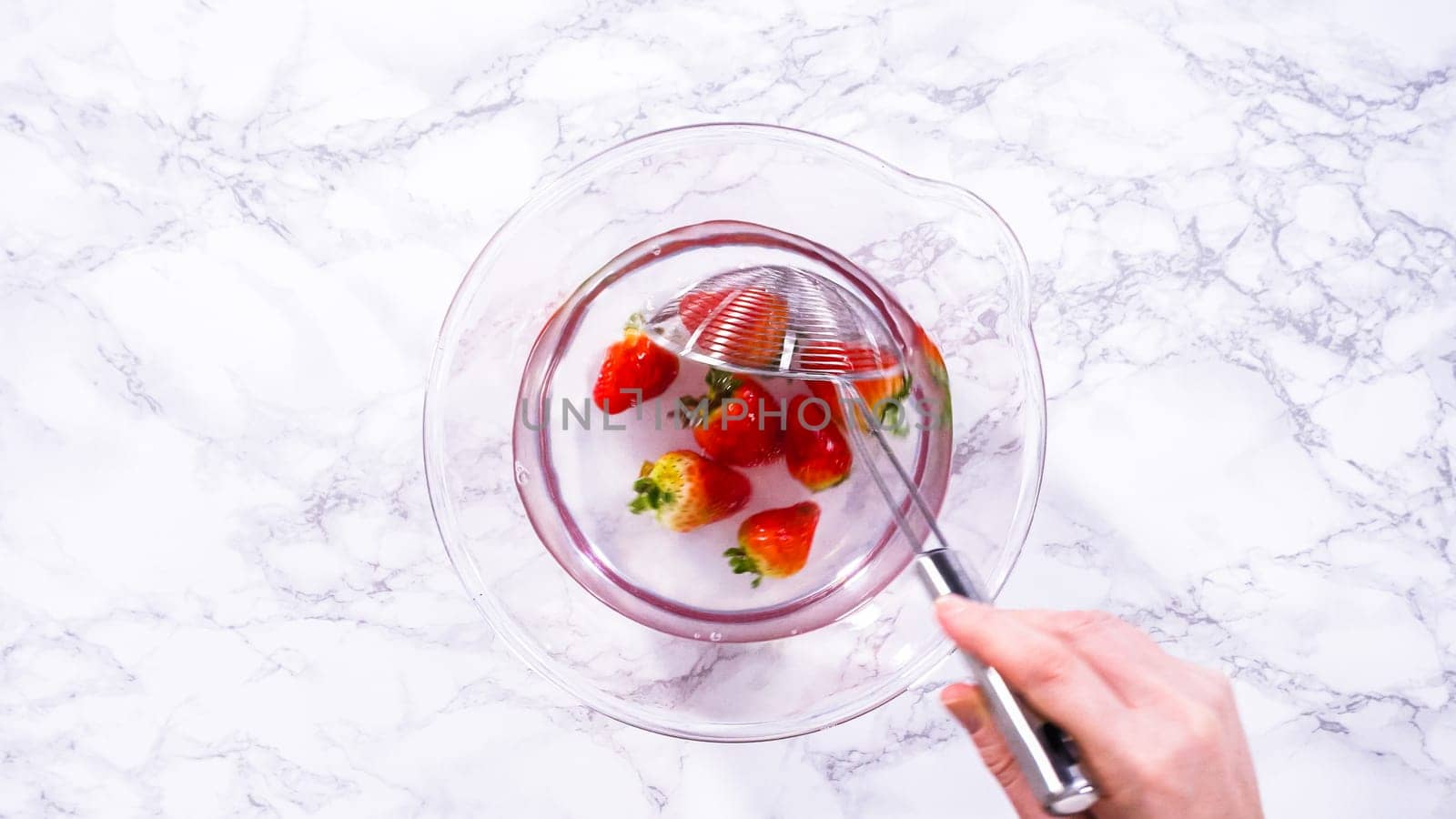 Flat lay. Ripe strawberries are submerged in water within a large glass mixing bowl, a step in washing the fruit to ensure cleanliness and longevity before storage or consumption.