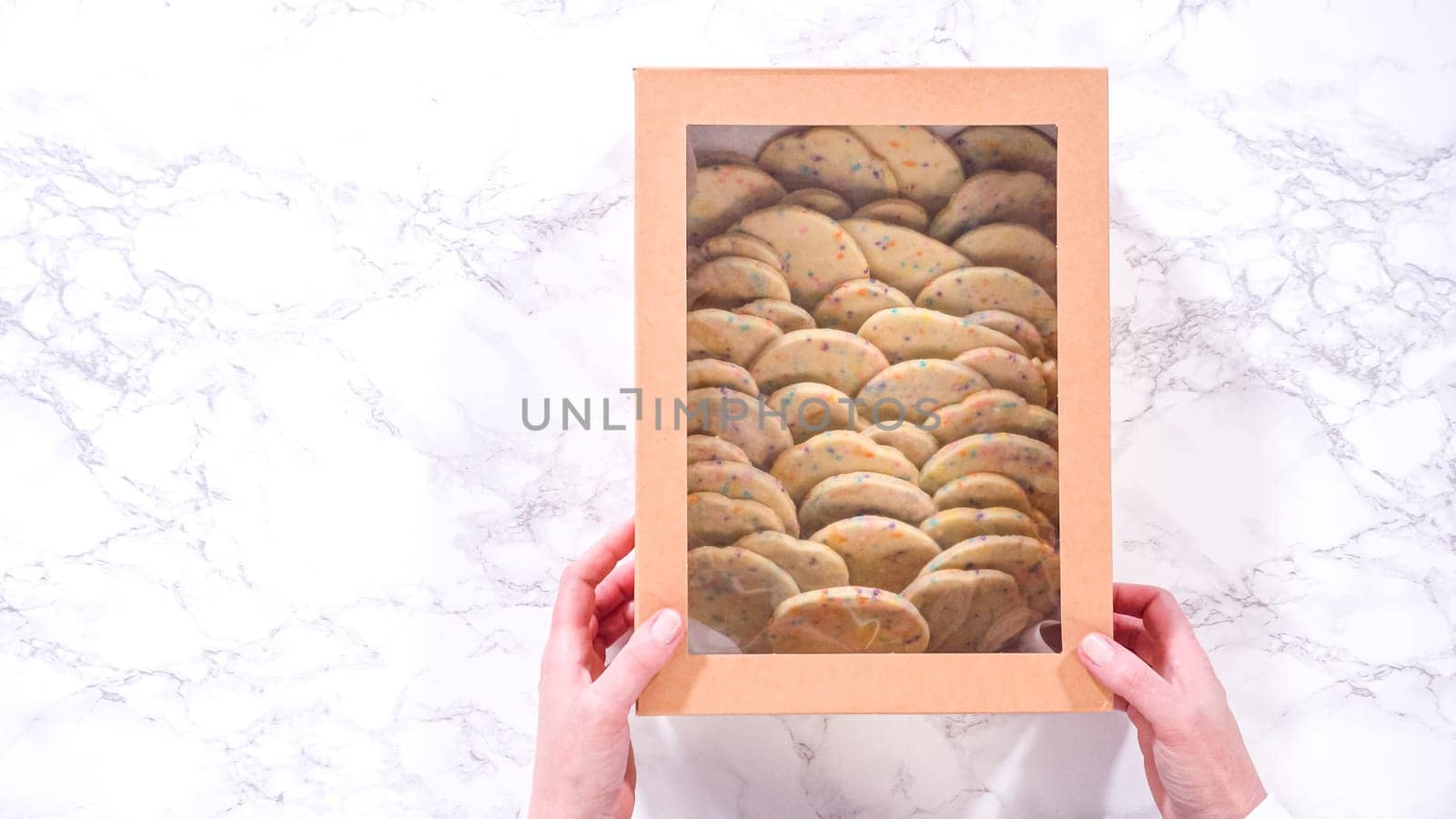 Flat lay. With precision, the woman is carefully arranging the sugar cookies, filled with dough-mixed sprinkles, into a rustic brown paper box.