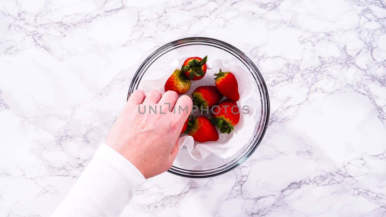 Flat lay. Strawberries, freshly washed and dried, are neatly stored in a glass bowl lined with a paper towel and securely covered with plastic wrap to maintain freshness.