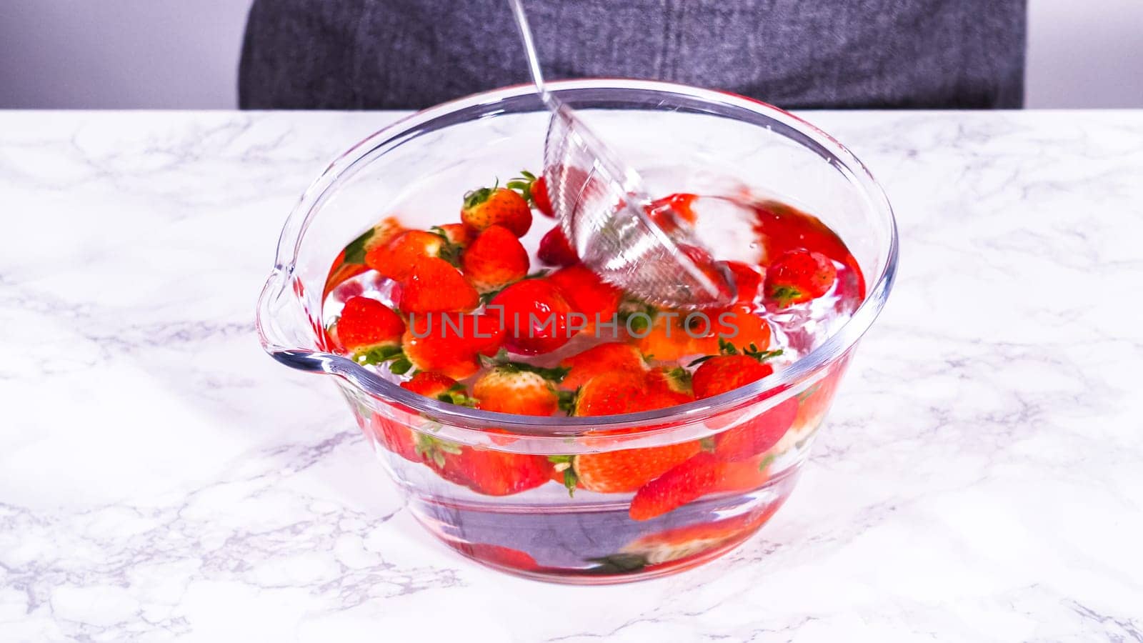 Ripe strawberries are submerged in water within a large glass mixing bowl, a step in washing the fruit to ensure cleanliness and longevity before storage or consumption.