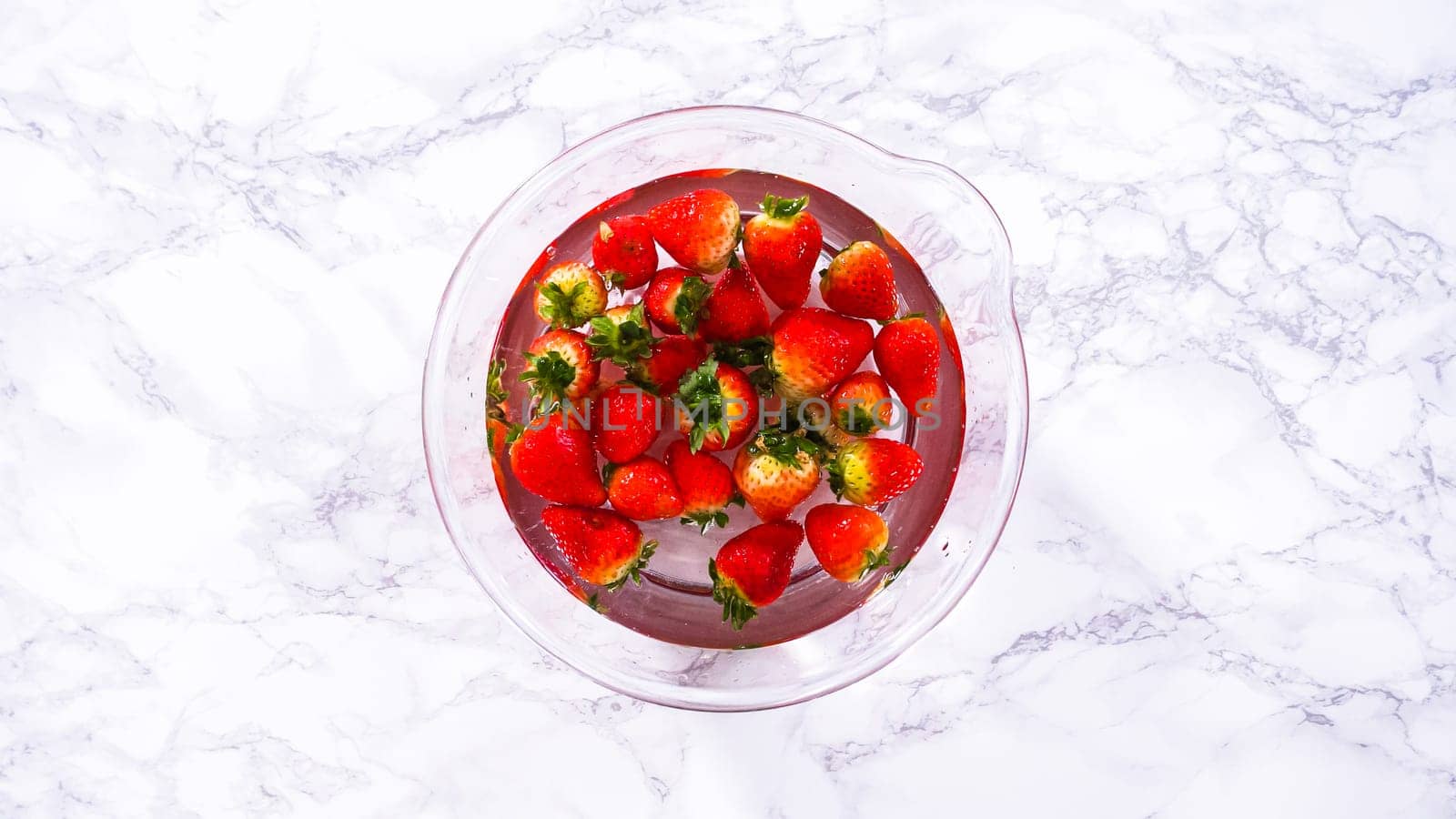 Flat lay. Ripe strawberries are submerged in water within a large glass mixing bowl, a step in washing the fruit to ensure cleanliness and longevity before storage or consumption.