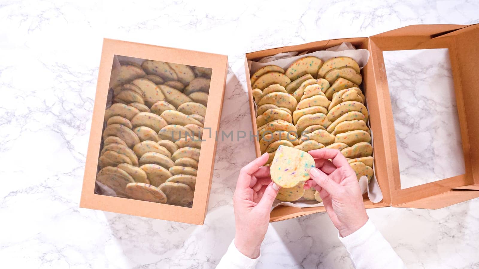 Flat lay. With precision, the woman is carefully arranging the sugar cookies, filled with dough-mixed sprinkles, into a rustic brown paper box.