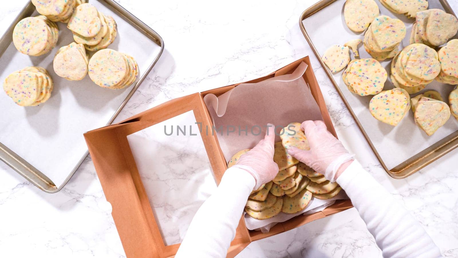 Flat lay. With precision, the woman is carefully arranging the sugar cookies, filled with dough-mixed sprinkles, into a rustic brown paper box.