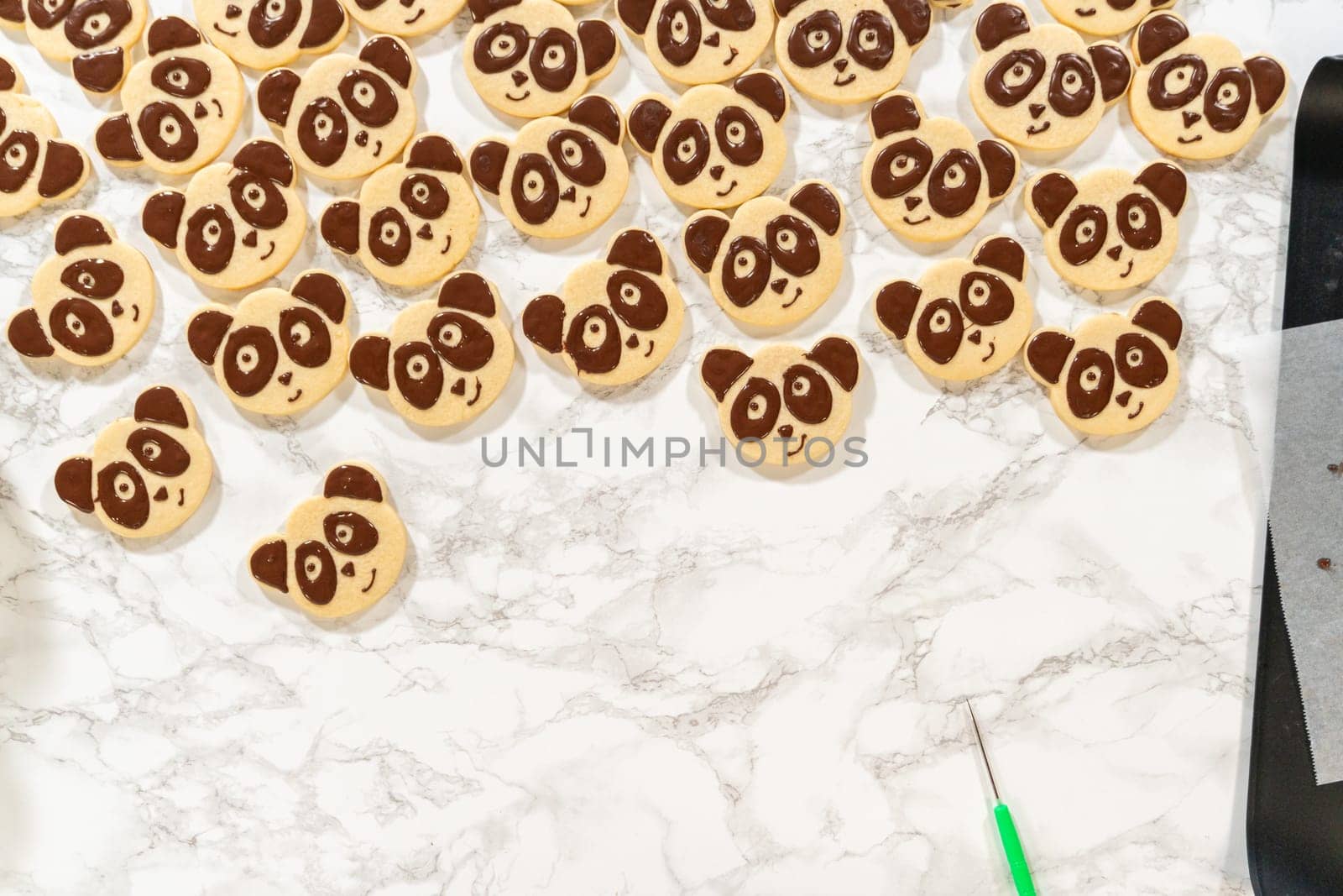 Overhead view of undecorated and panda-face decorated shortbread cookies, with two piping bags filled with chocolate royal icing, arranged on a marble surface.