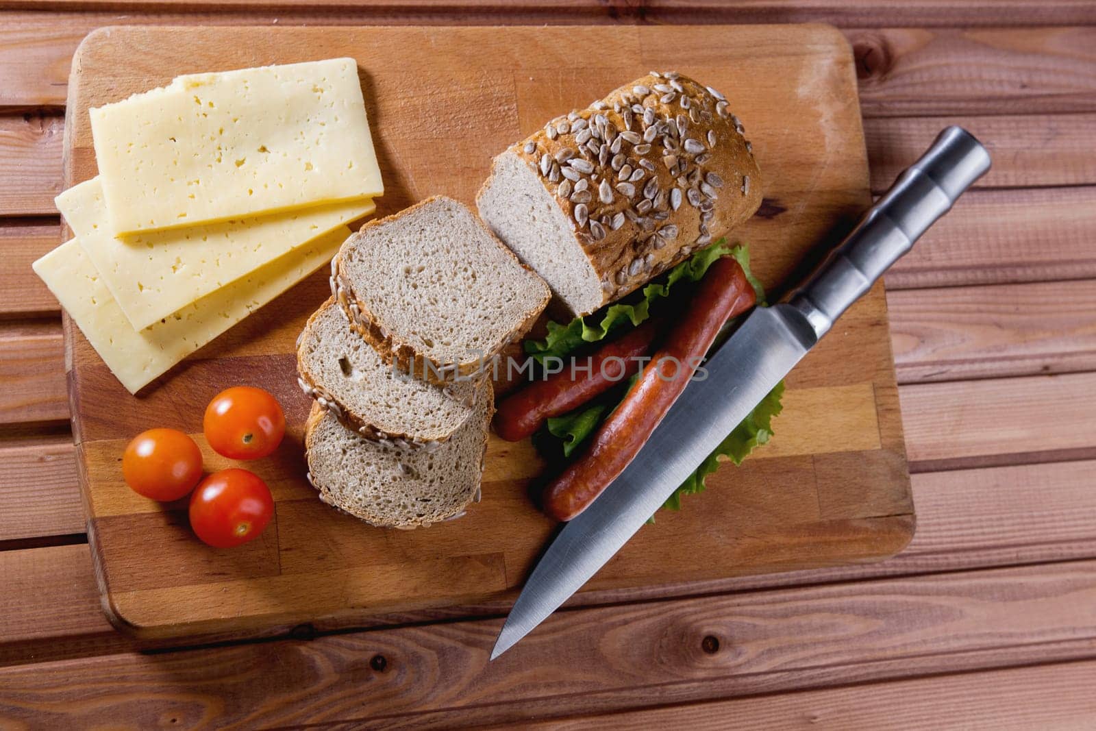 Fried sausages on green salad, bread, cherry tomatoes and cheese on wooden board, close up - top view