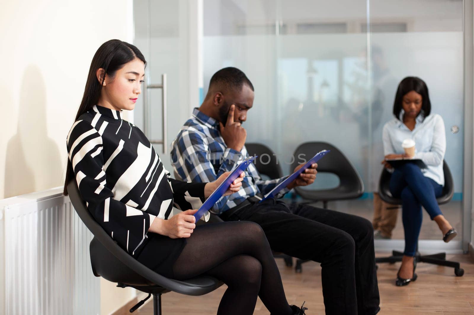 Multiethnic job candidates gather in office lobby for HR meeting, reviewing recruitment documents and holding resumes. Young applicants wait in line for interviews, reading hiring materials.