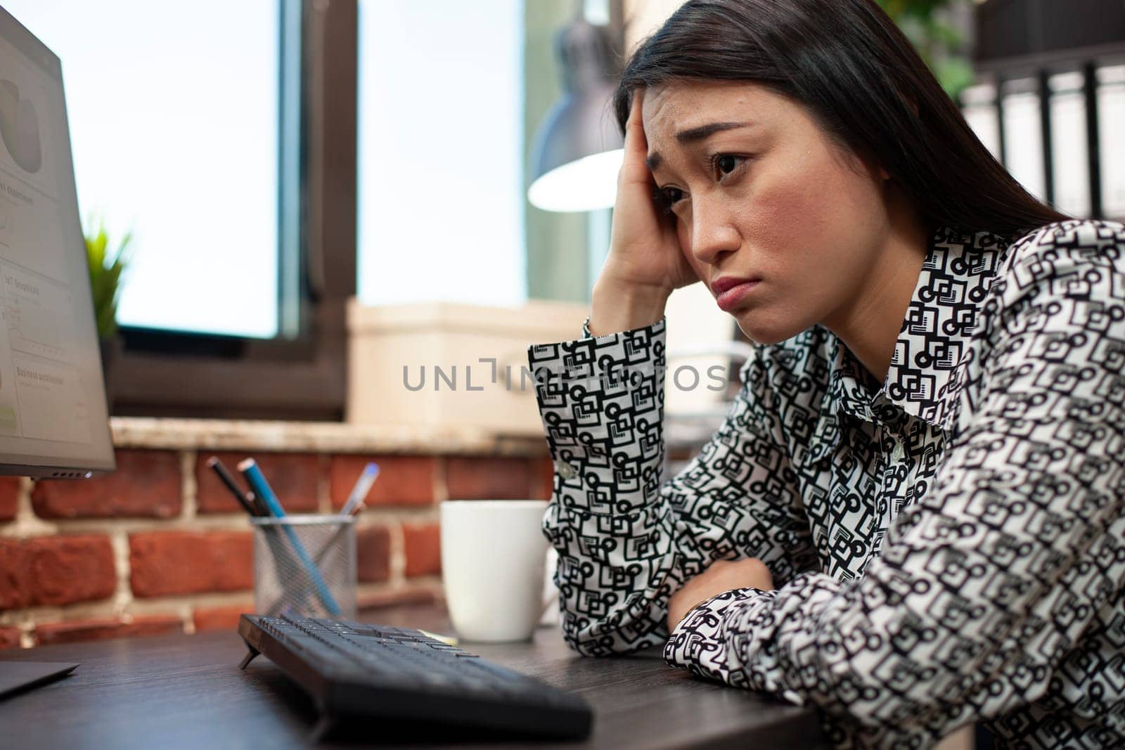 Closeup of exhausted entrepreneur resting her face on her palm at desk after a long workday. Businesswoman with hand on forehead trying to stay alert while working on computer in a startup office.