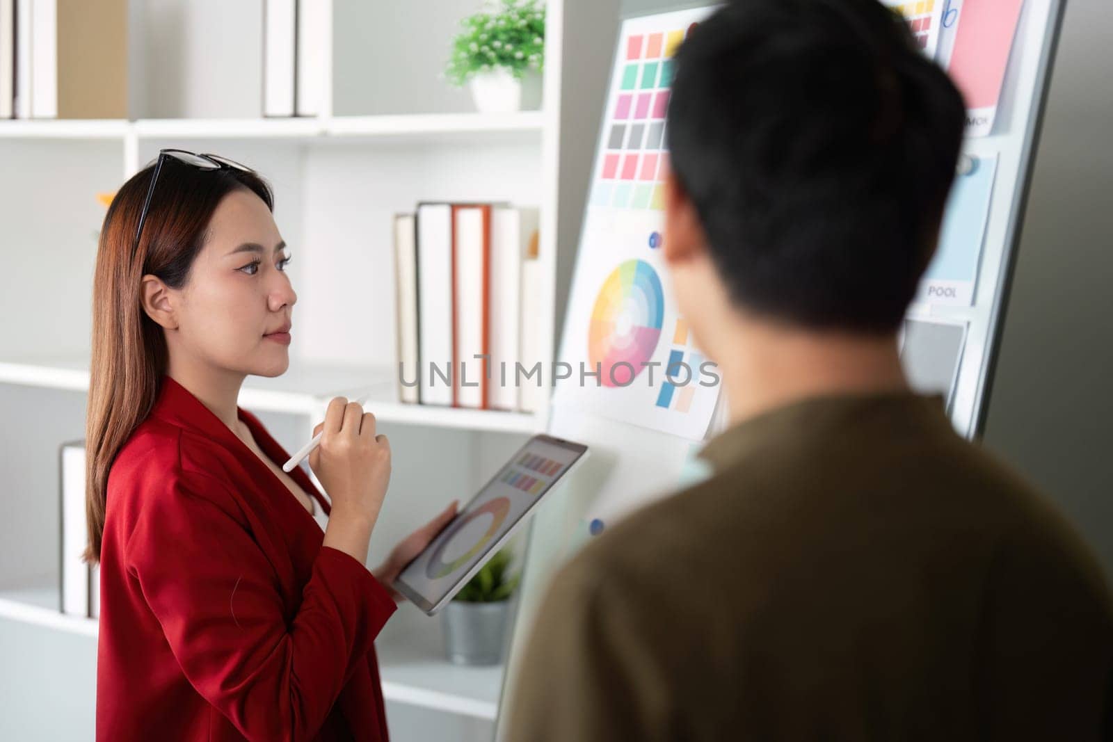 In a dynamic office setting, a female designer evaluates her color wheel on a digital tablet, engaging creatively with her colleague.