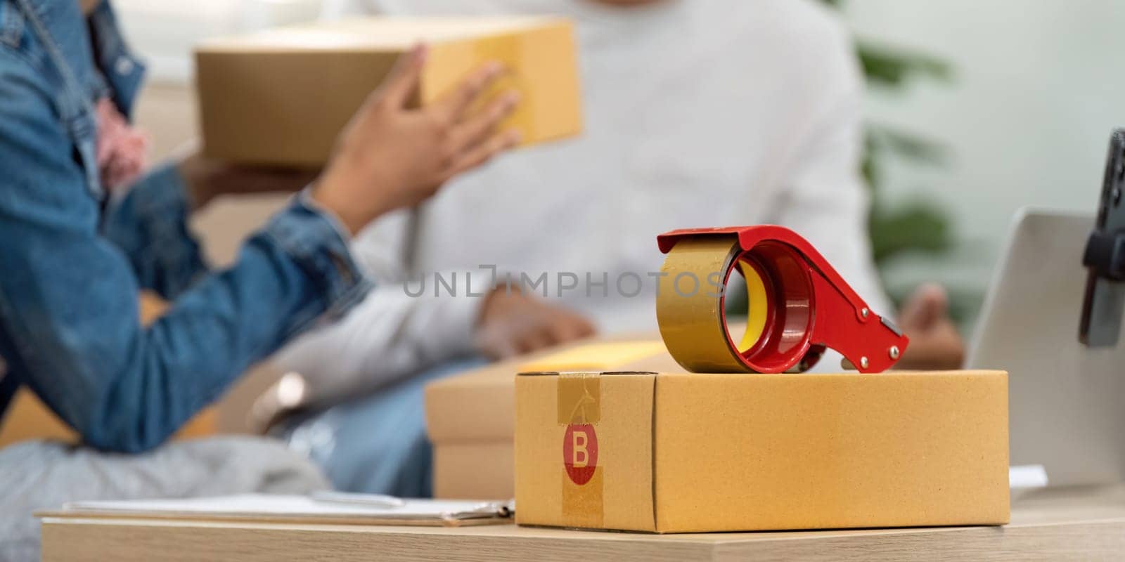 Essentials for shipping, including boxes and tape, are set up on a table as a couple prepares their entrepreneurial shipment for their online business.