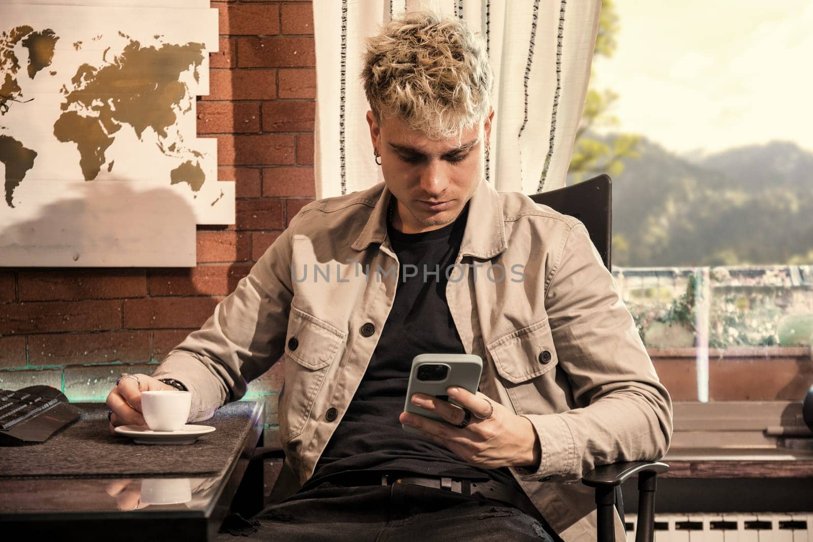 A young man with attractive blond hair and captivating blue eyes sits in a comfortable cafe, savoring a cup of coffee while focused on his smartphone. Natural light streams in through the window, enhancing the warm ambiance.