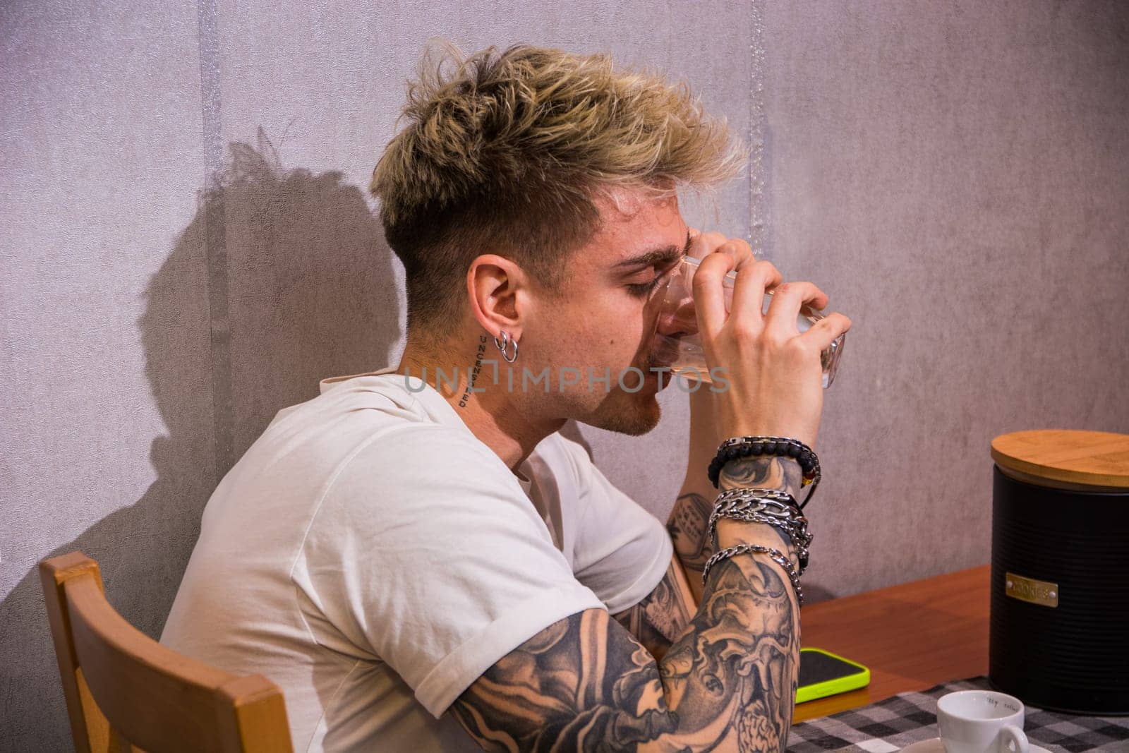A young man with blond hair and blue eyes sits at a table, sipping a drink. Surrounded by a relaxed atmosphere, he showcases his tattoos and casual style while enjoying his moment.